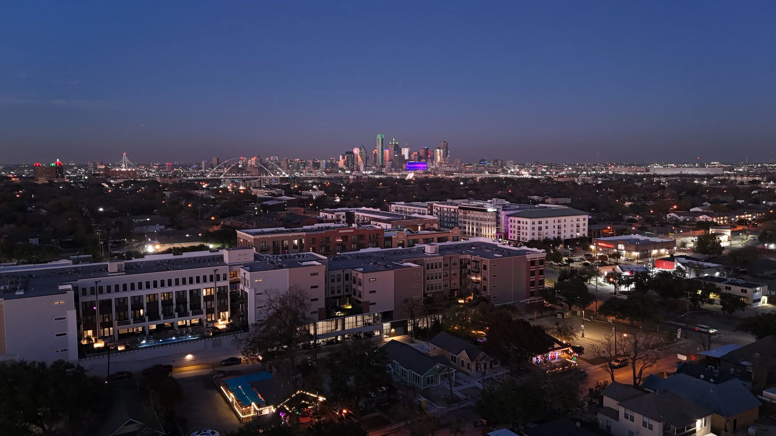 City skyline at dusk beyond low-rise buildings, with illuminated streets and an arched bridge.