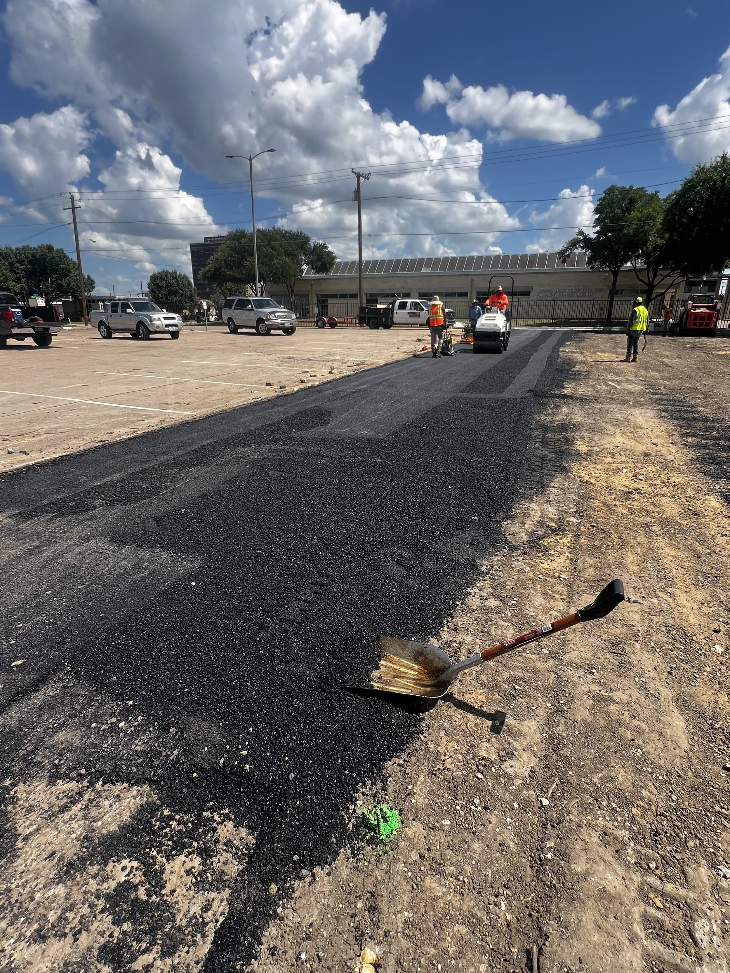 Crew members and a roller working along a strip of asphalt beside a prepared parking area.