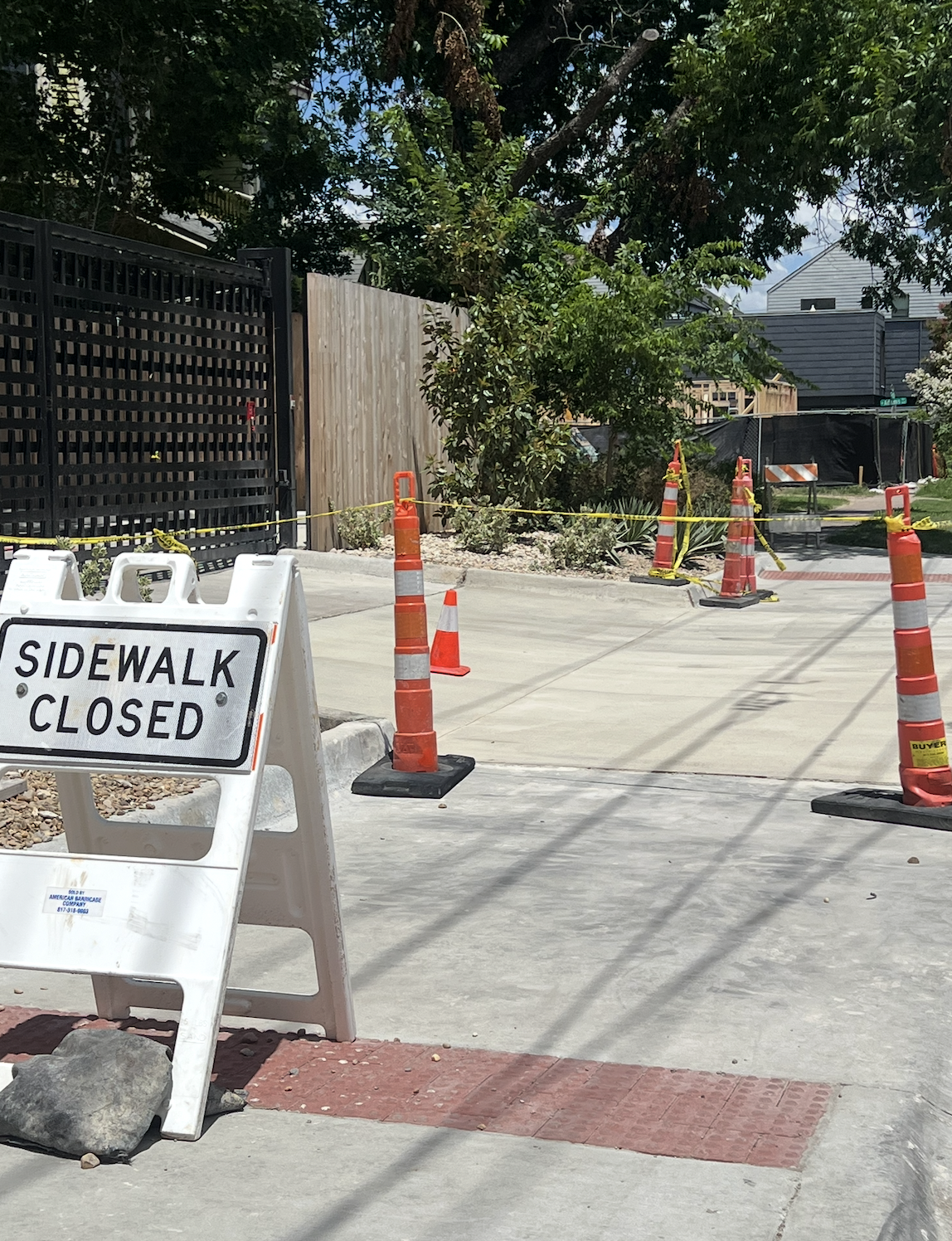 Repaired concrete sidewalk beside a black gate, with orange cones and a sidewalk closure sign.