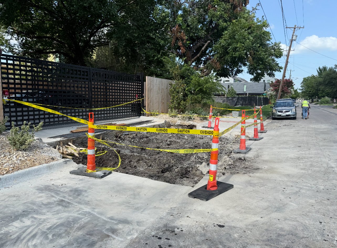 Excavated sidewalk work area marked with orange cones and yellow caution tape.
