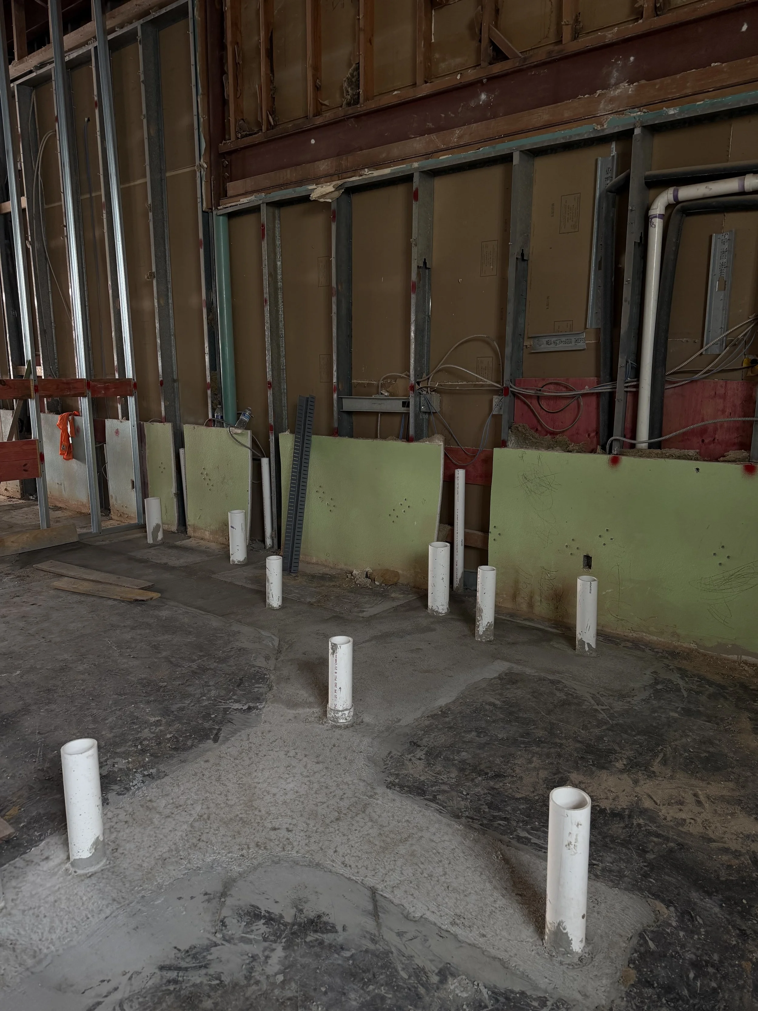 Unfinished interior with metal wall framing, horizontal wood backing, and white pipe stub-outs rising from the floor.