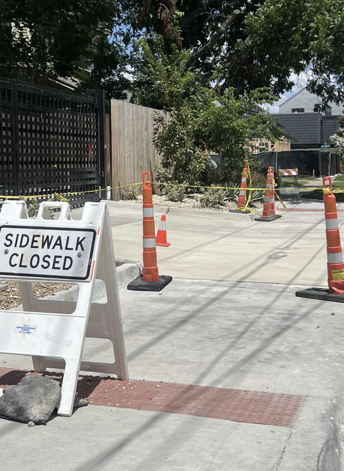 Repaired concrete sidewalk beside a black gate, with orange cones and a sidewalk closure sign.