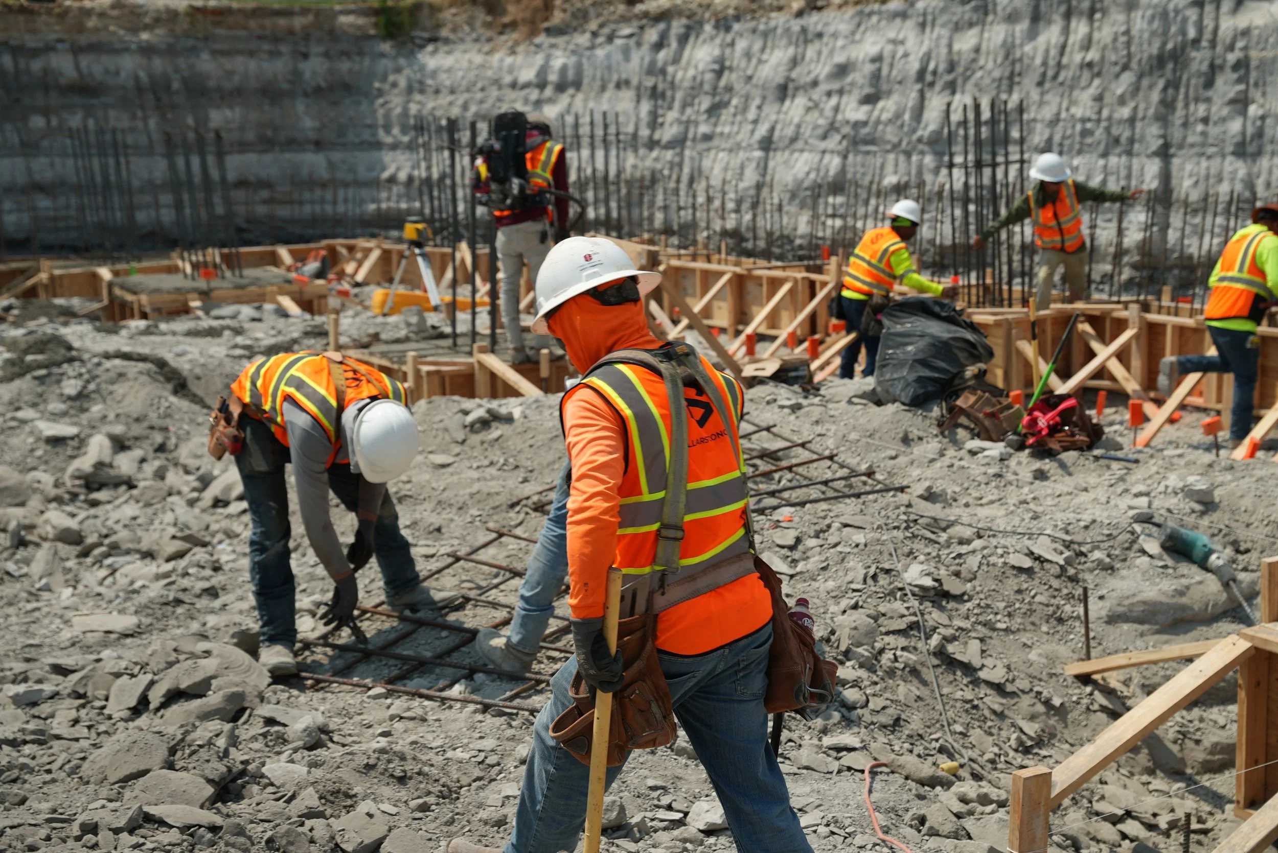 Crew members wearing hard hats and safety vests working among steel reinforcement, timber forms, and excavated ground.