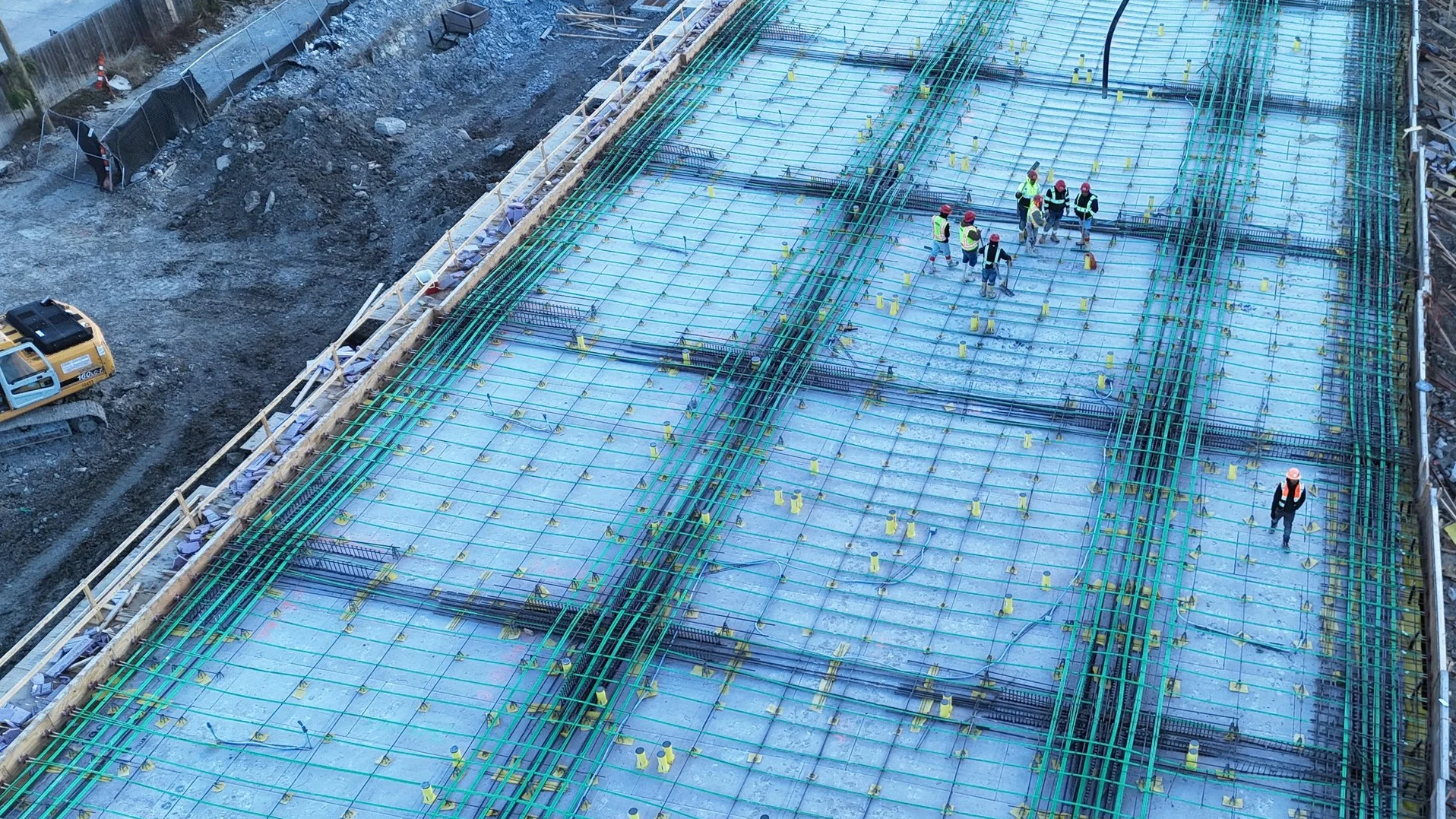 Aerial view of workers standing among steel reinforcement grids over a blue membrane at a construction site.