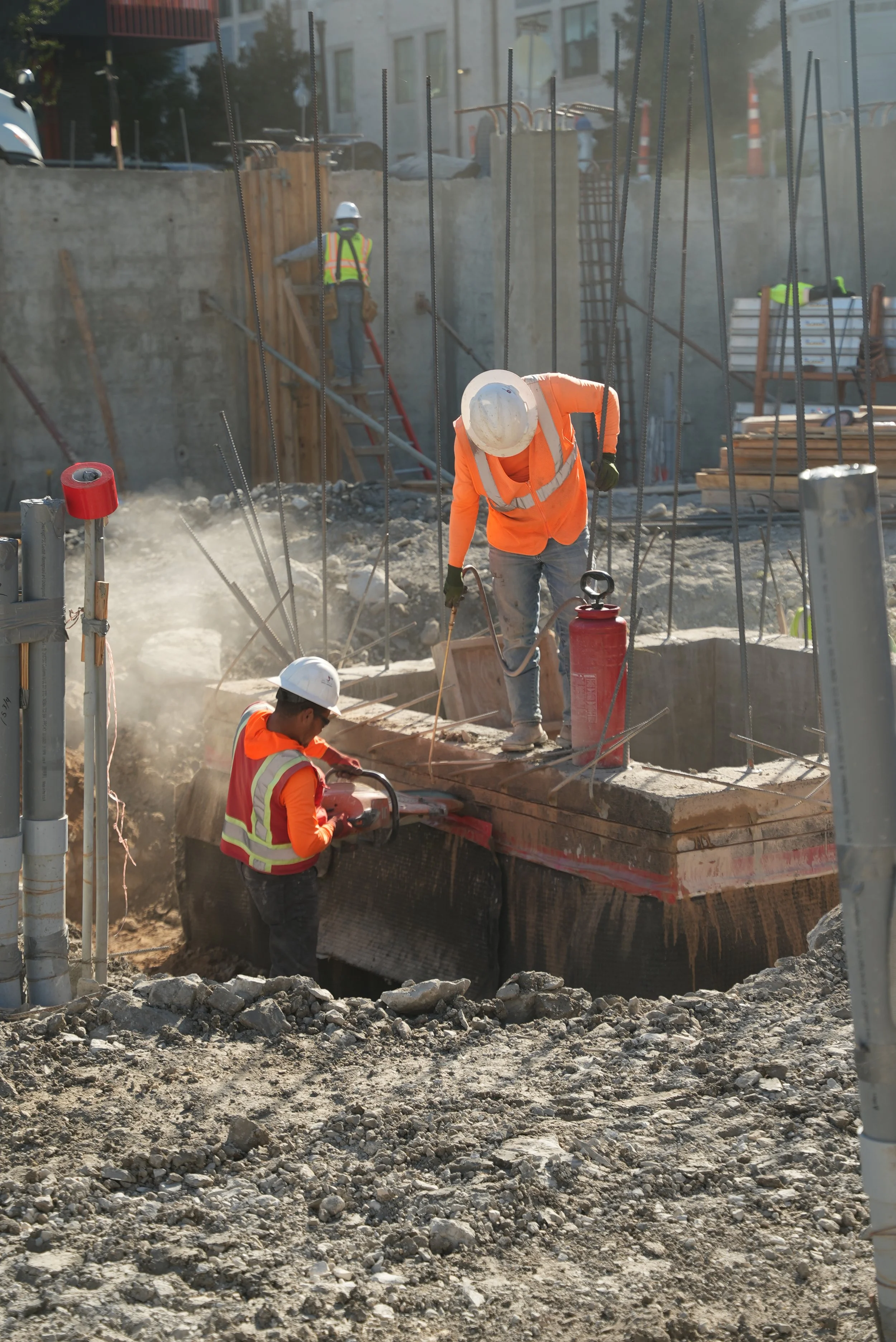 Workers in orange safety vests working beside timber forms and vertical reinforcement at a foundation work area.