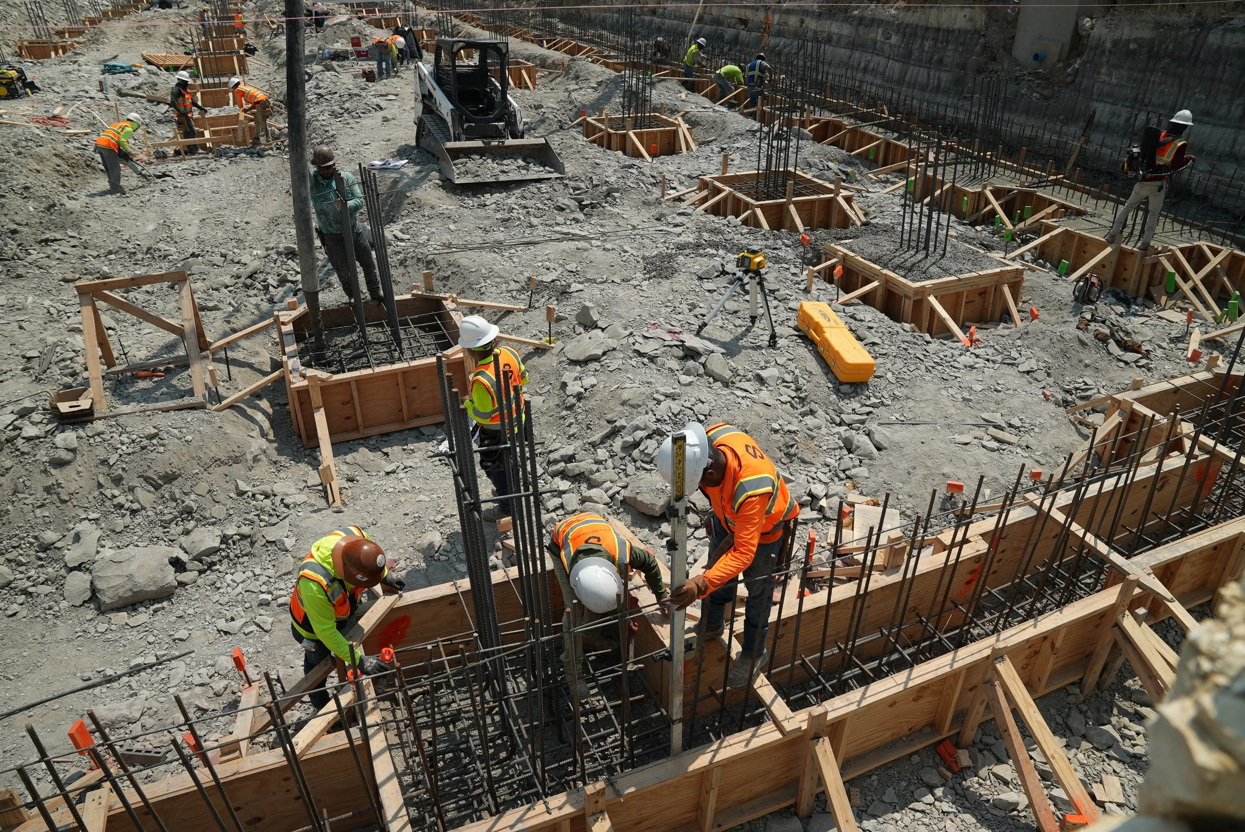 Crew working around reinforcing cages and timber foundation forms at an excavated construction site.