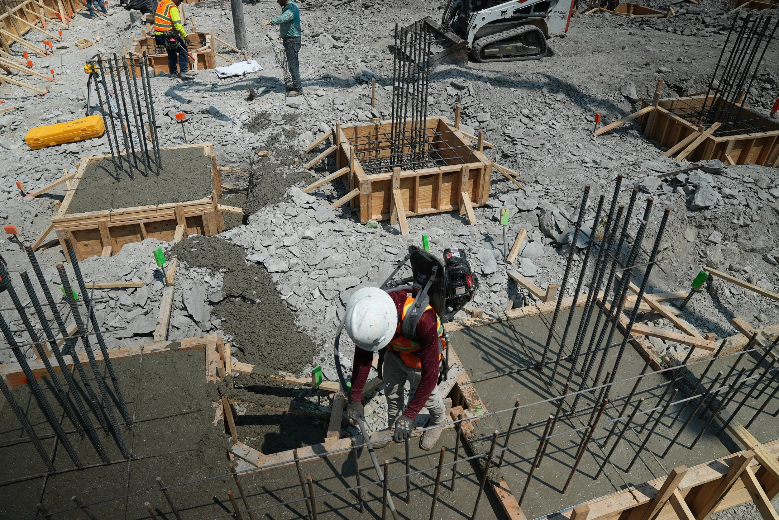 Worker tending fresh concrete around vertical reinforcing bars inside timber foundation forms.