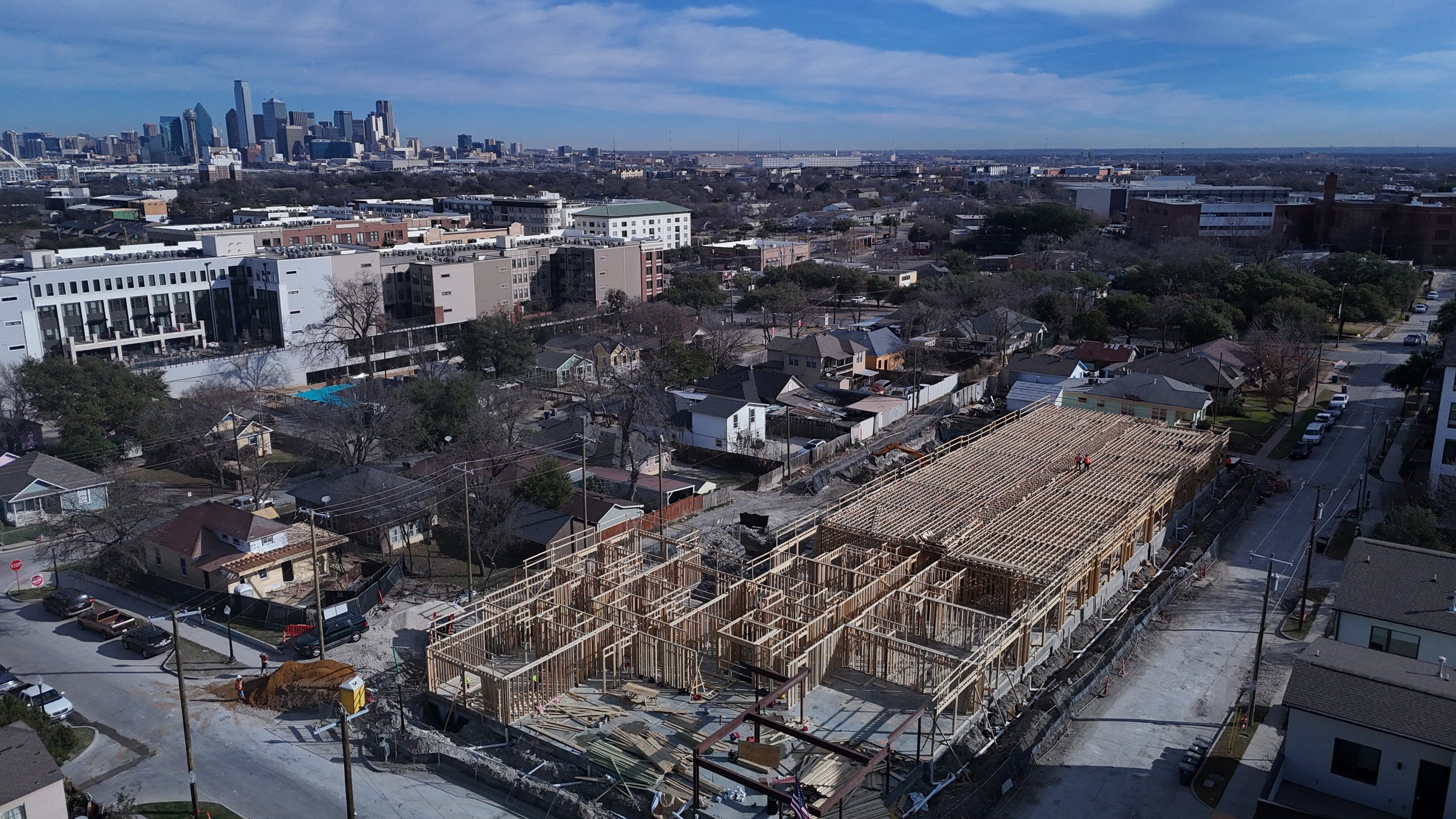 Aerial view of a wood-framed construction site with surrounding streets and a city skyline in the distance.