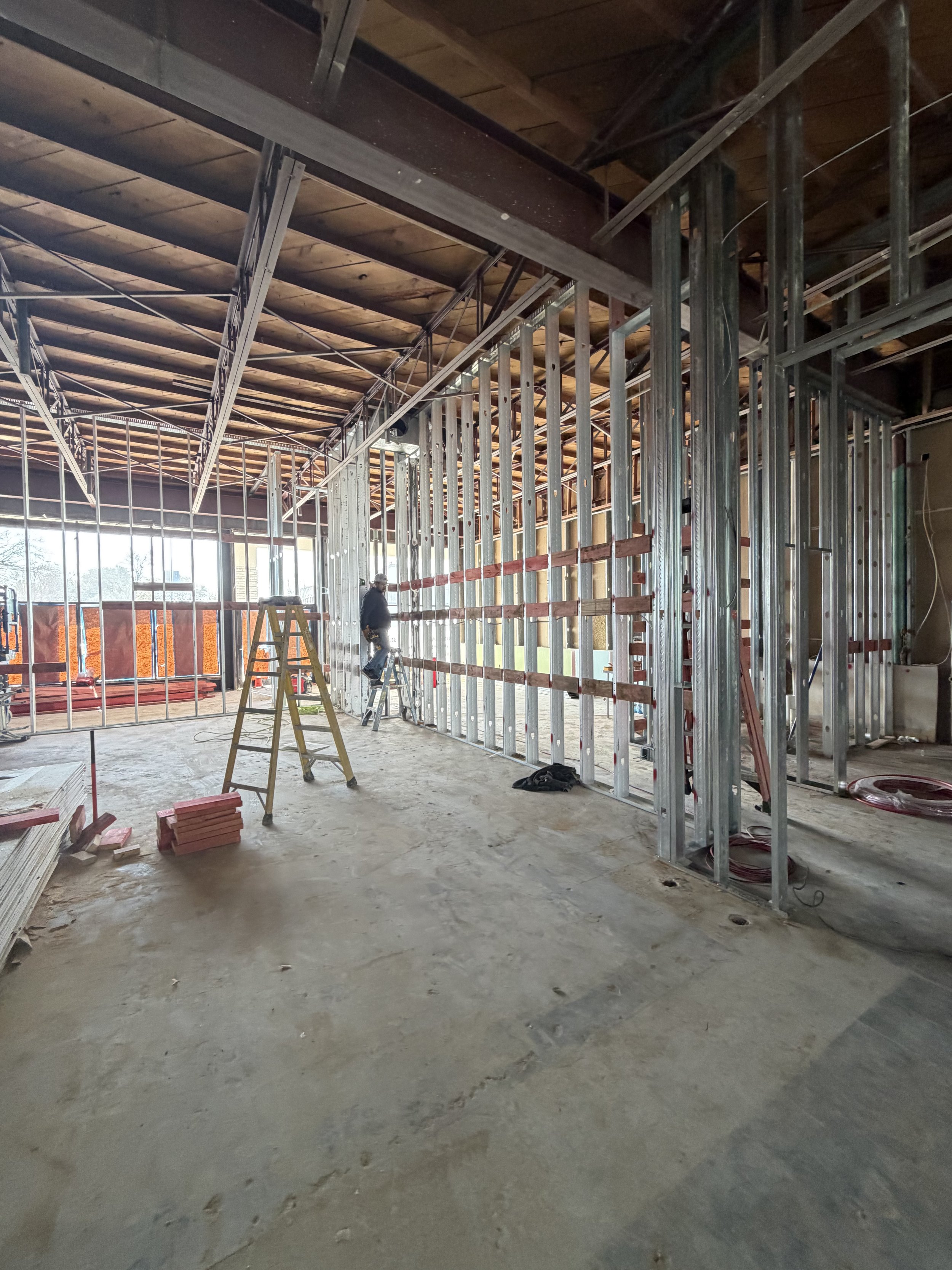 Exposed ceiling structure and metal wall framing inside a commercial interior, with a ladder and construction materials.