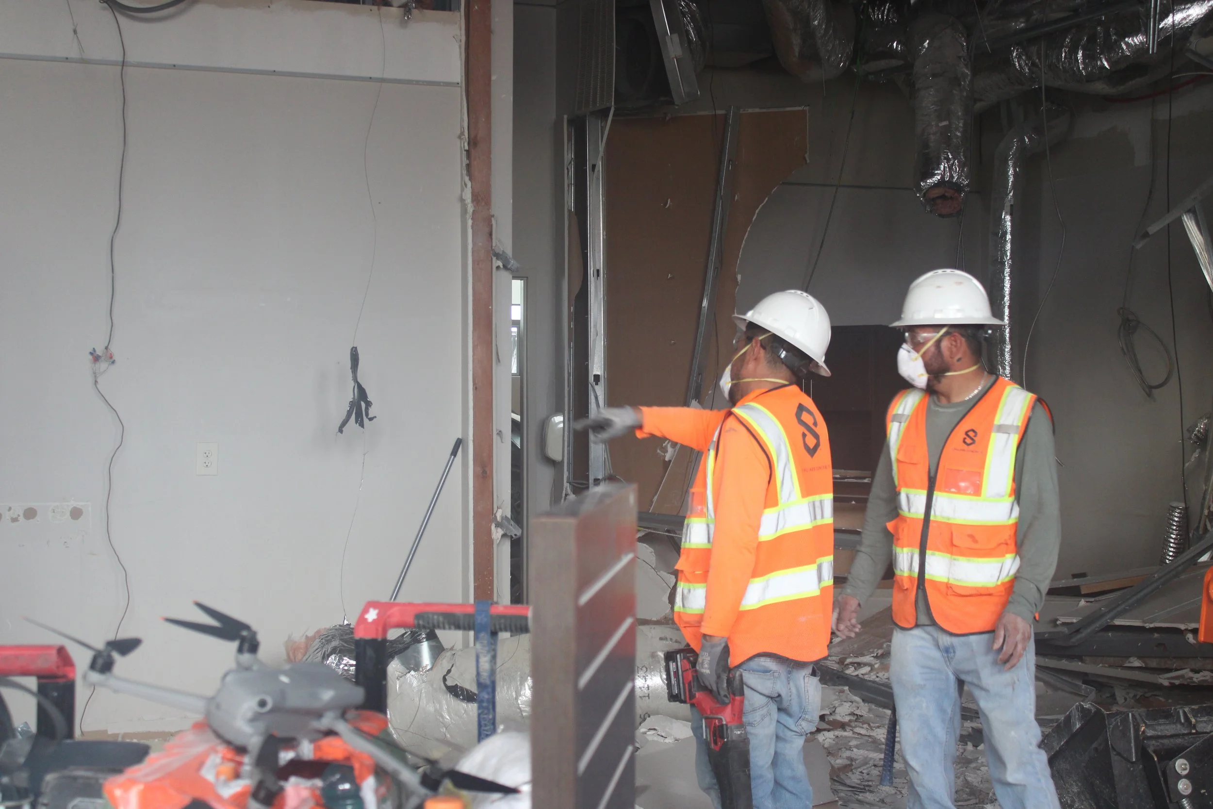 Two workers wearing hard hats and orange safety vests beside an unfinished interior wall.