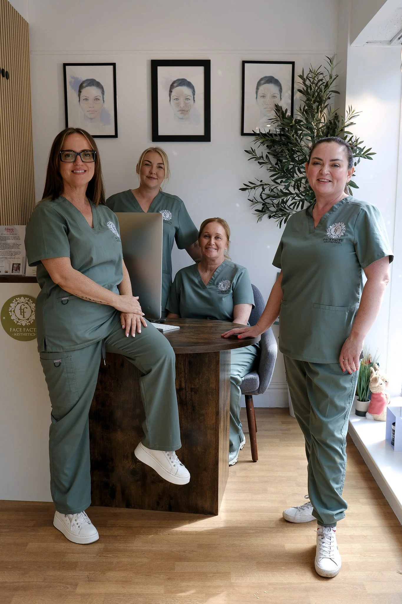 Four women wearing green medical scrubs in a professional setting, with framed portraits behind them and a large plant nearby.