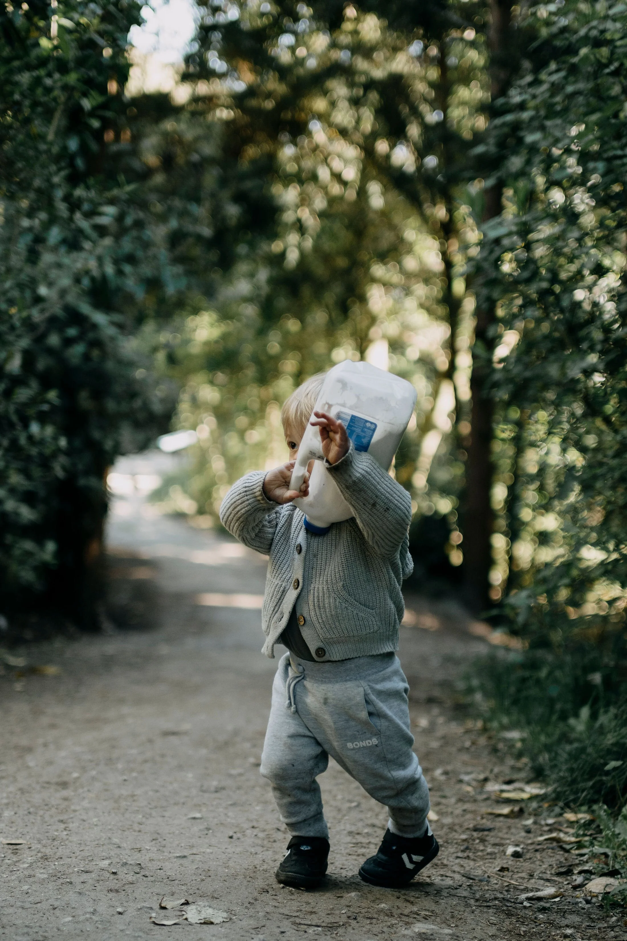 A young boy with blonde hair, wearing a grey sweater, grey pants, and black shoes, walking on a dirt pathway in a wooded area with trees and sunlight in the background.