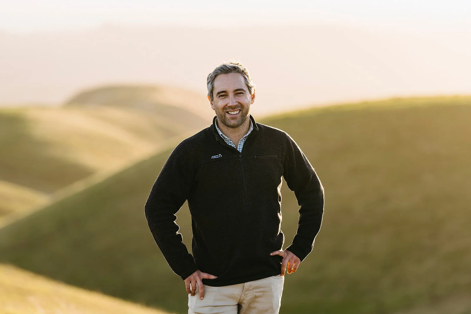 A smiling man with gray hair and beard standing outdoors on a grassy hill during golden hour, wearing a black zip-up fleece and beige pants.