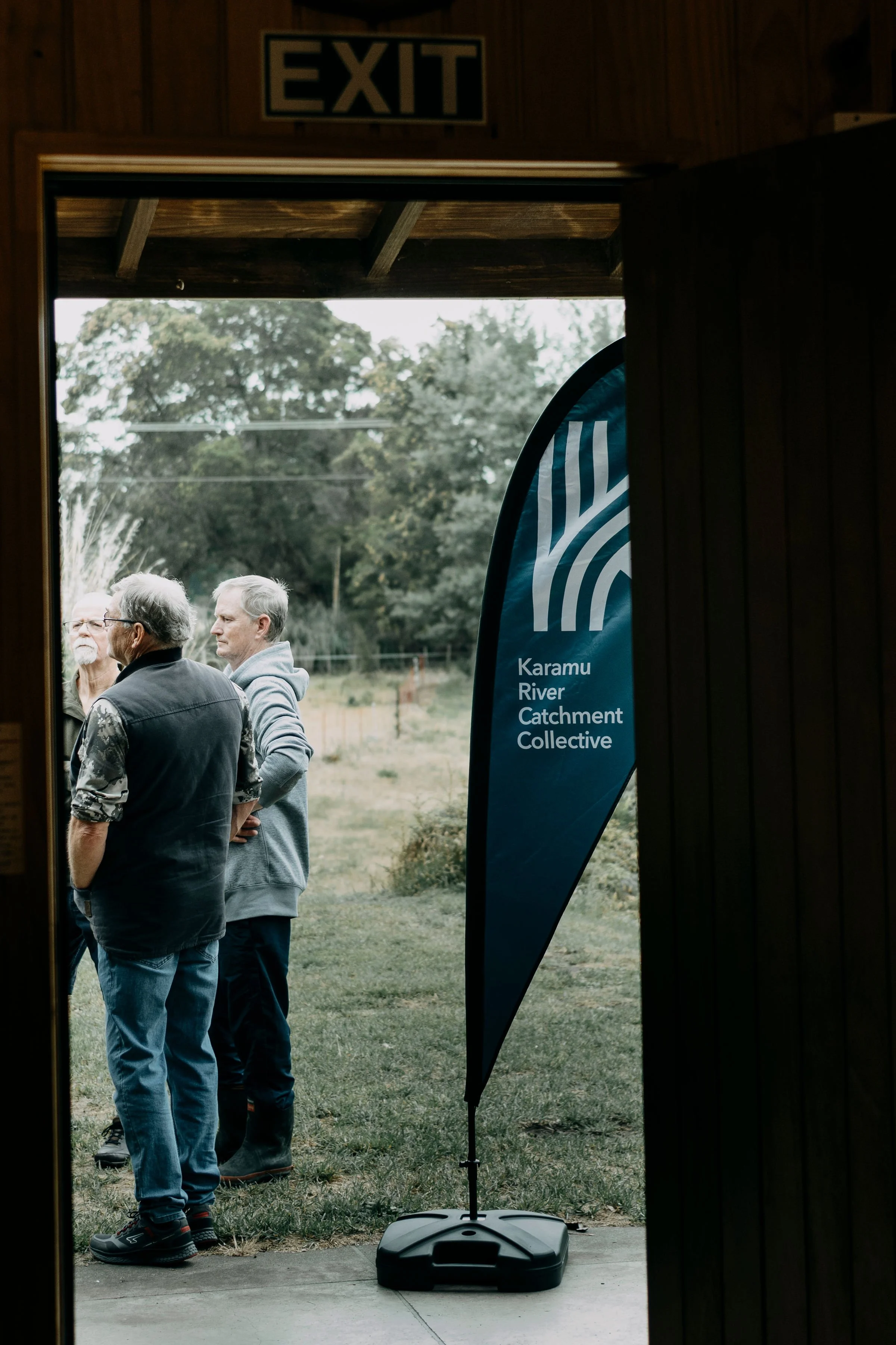 A group of people standing outdoors near trees, viewed from inside a building through an open doorway. There is a blue flag/banner with white text that reads 'Karamu River Catchment Collective.' An exit sign is visible at the top.