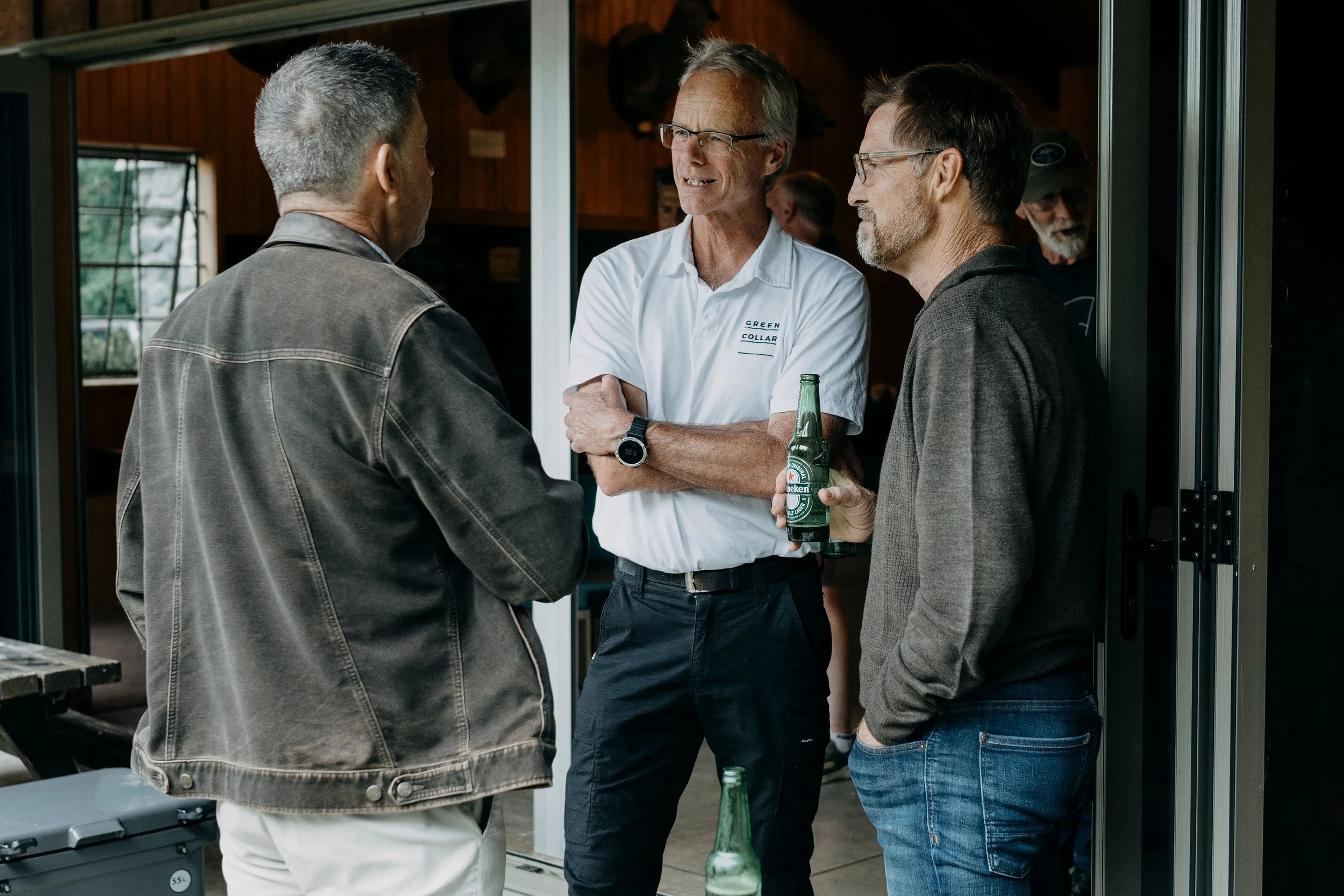 Three men are having a conversation outside a building, with one holding a Heineken beer bottle. The man in the middle wears glasses and a white shirt, while the other two are dressed casually in dark jackets.