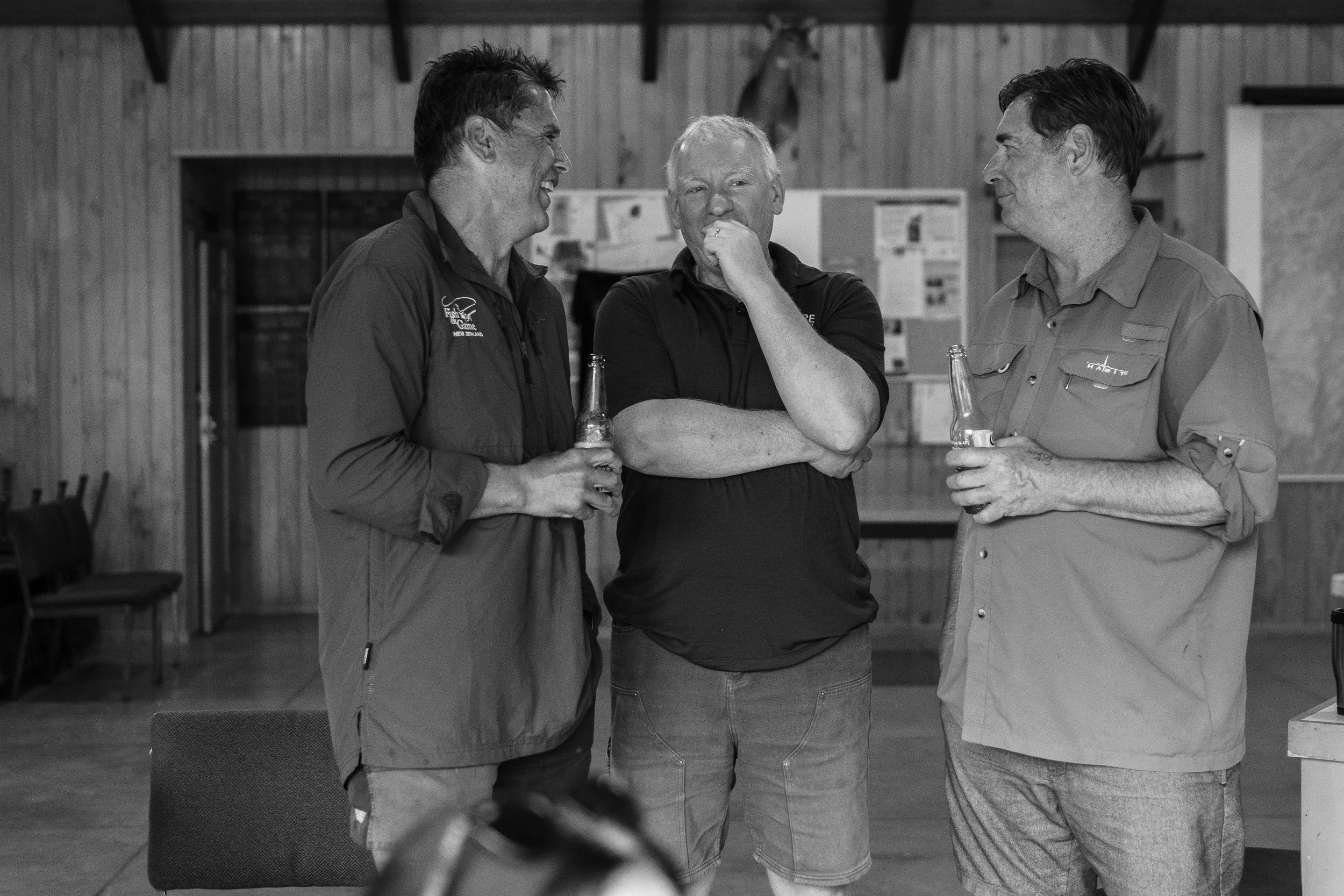 Three men are talking and laughing indoors in a wooden room, each holding a bottle. The man on the left wears a jacket with writing that says 'Fish' and 'New Zealand'; the man in the middle has light hair and wears a dark shirt; the man on the right has dark hair, a tattoo on his hand, and wears a light button-up shirt with a patch that says 'WABIT'.