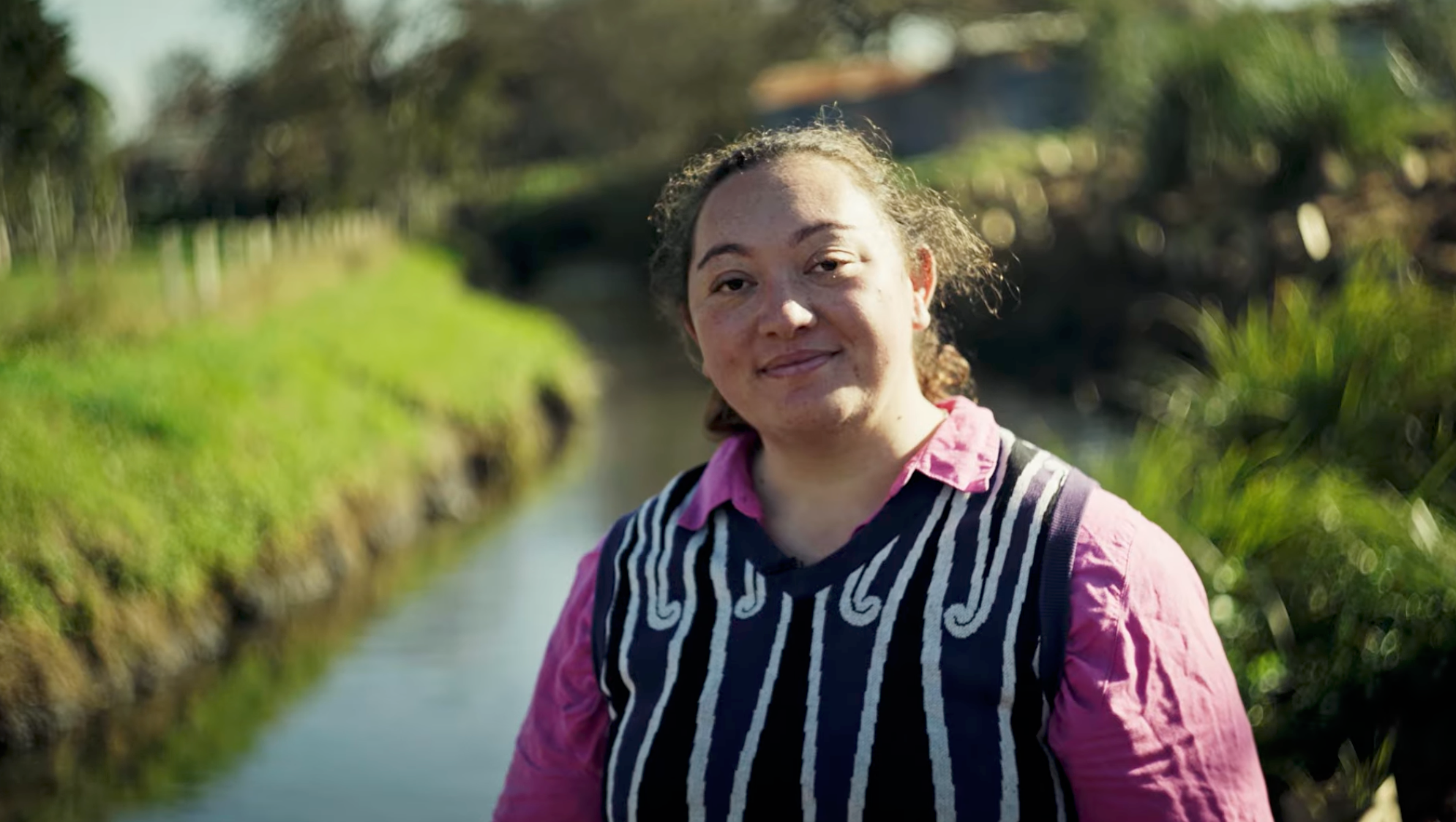 A person with curly hair and a pink shirt wearing a striped vest, standing outdoors near a waterway with lush green trees and grass in the background on a sunny day.