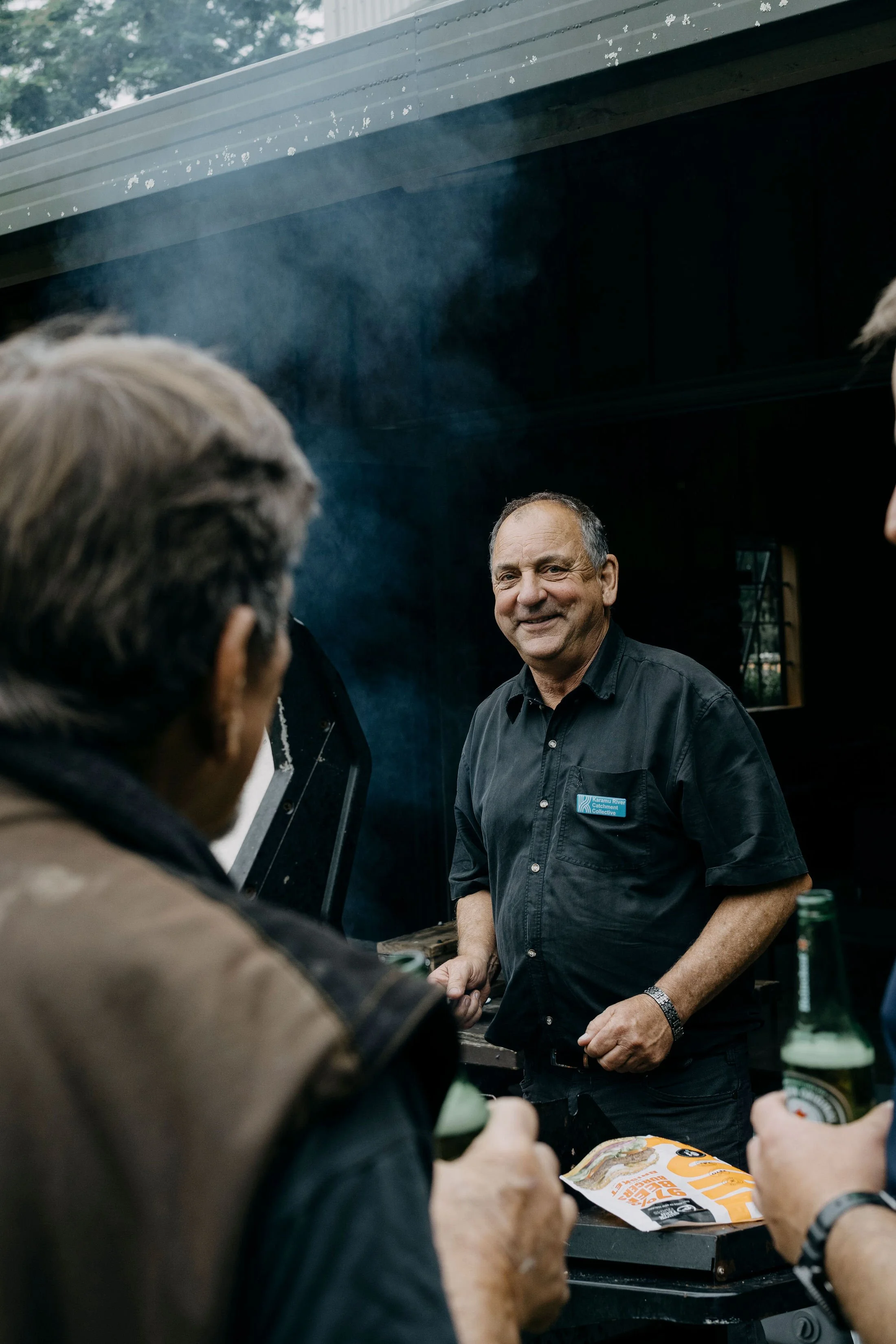 A man with a gray beard and wearing a dark shirt is smiling and shaking hands with someone during an outdoor gathering, with people in the foreground and trees in the background.