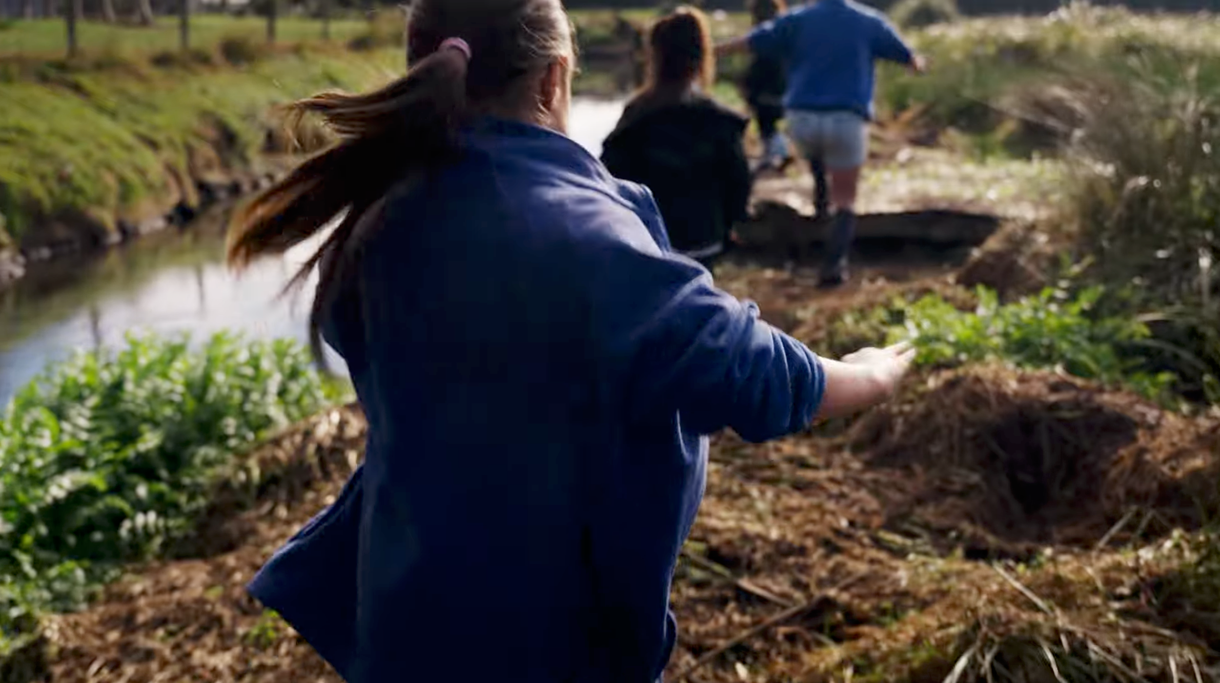 Children run along a bank of a small creek or pond in a green outdoor area during daytime.