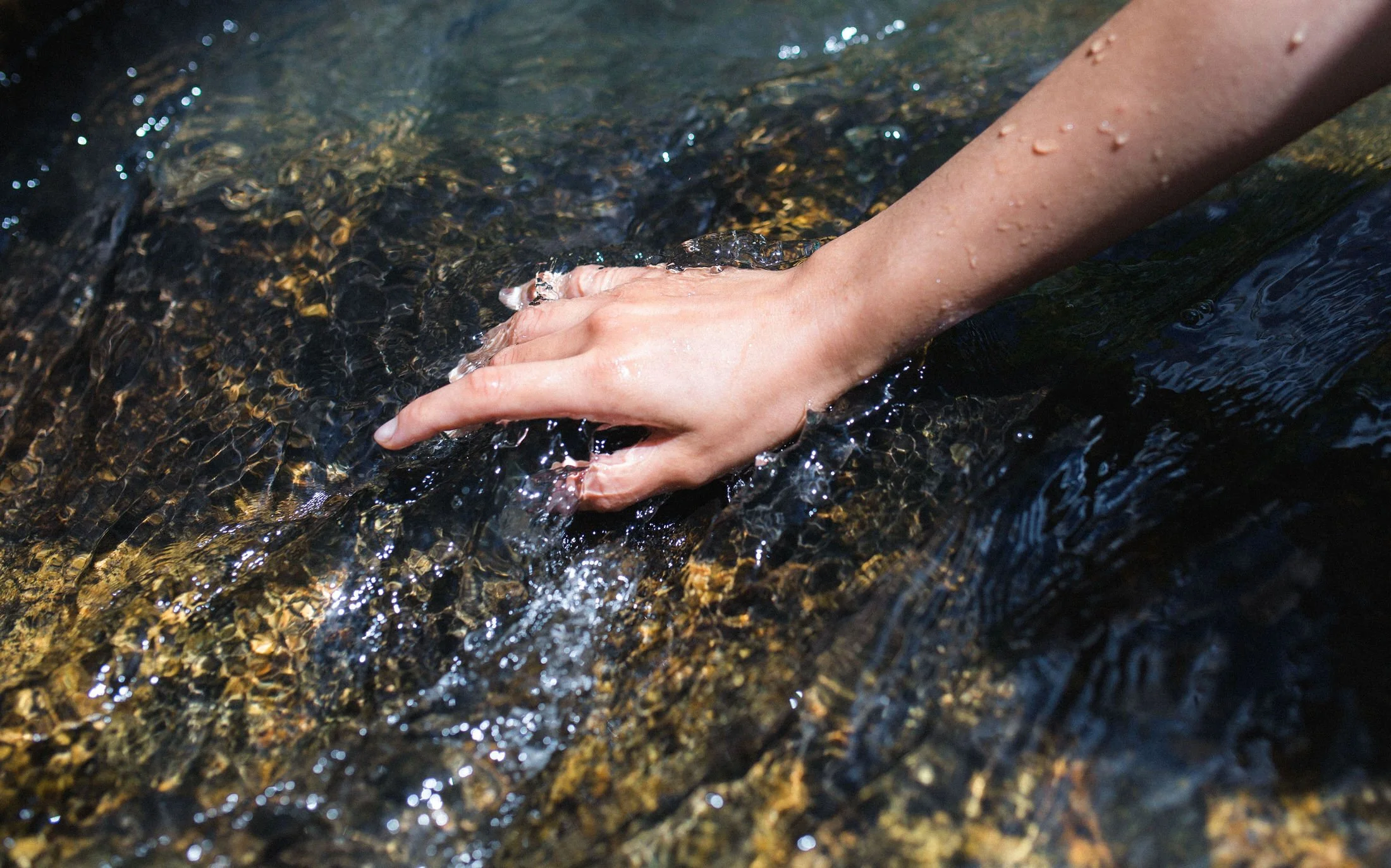 A person’s hand submerged in a clear, shallow stream with visible pebbles on the bottom, water flowing over the hand.