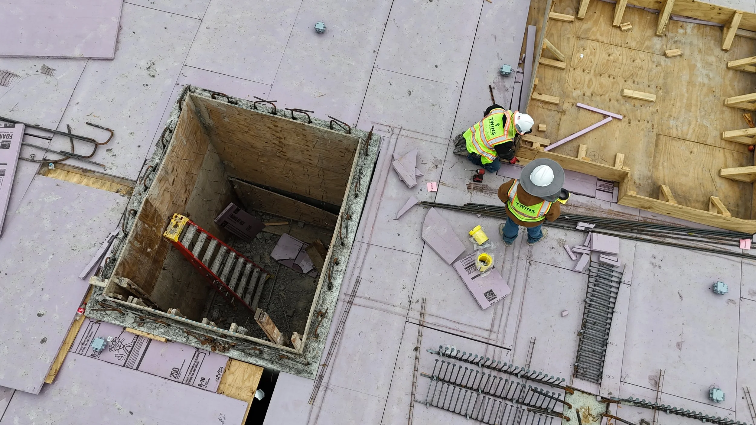 Overhead view of two workers wearing safety vests beside timber formwork and steel reinforcement on a construction deck.