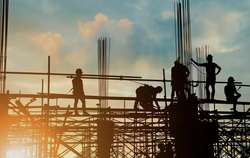Silhouetted construction worker on scaffolding beside vertical reinforcing bars at sunset.