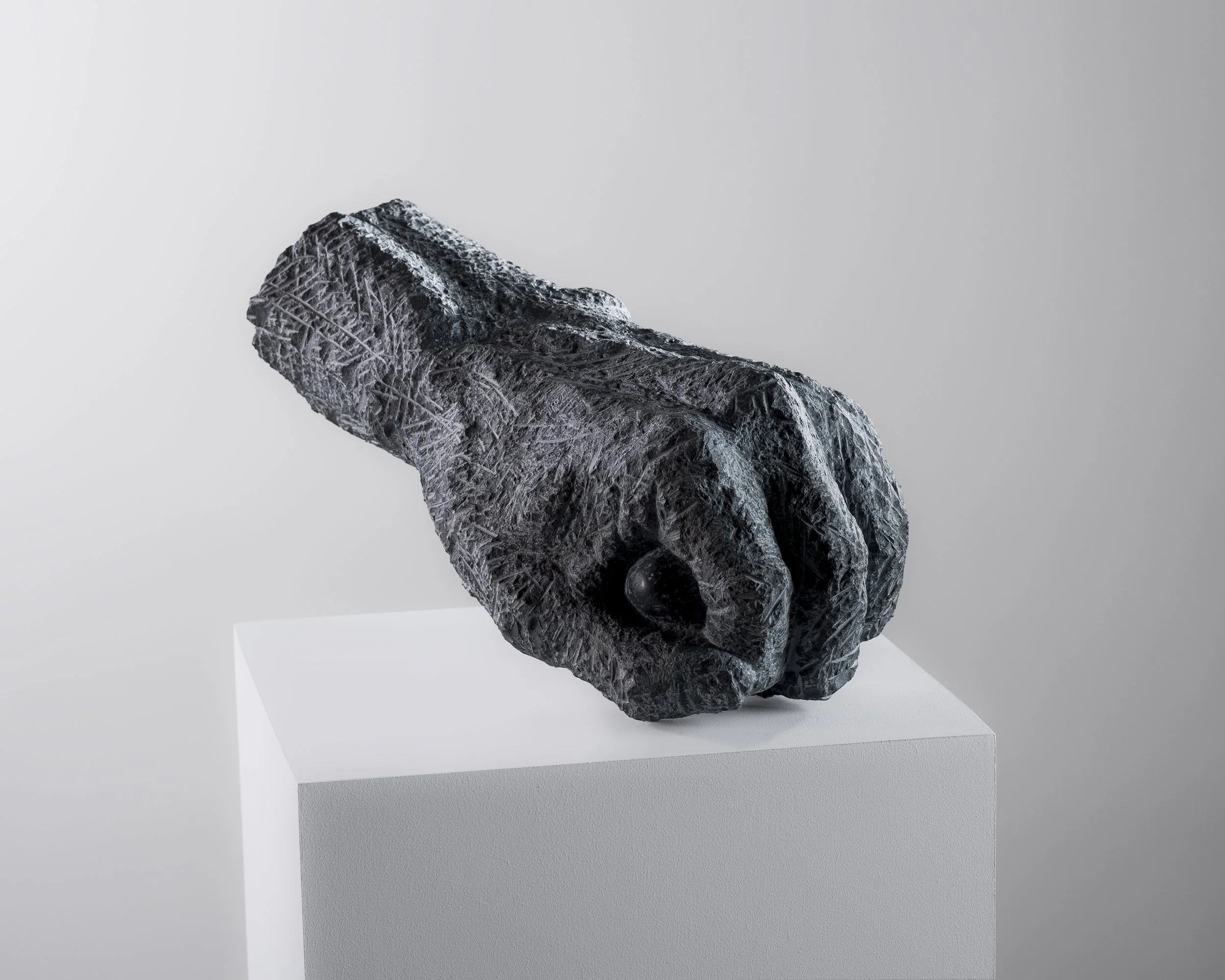 Sculpture en pierre sombre avec une surface texturée et des détails complexes, exposée sur une plateforme blanche dans un environnement d'exposition.