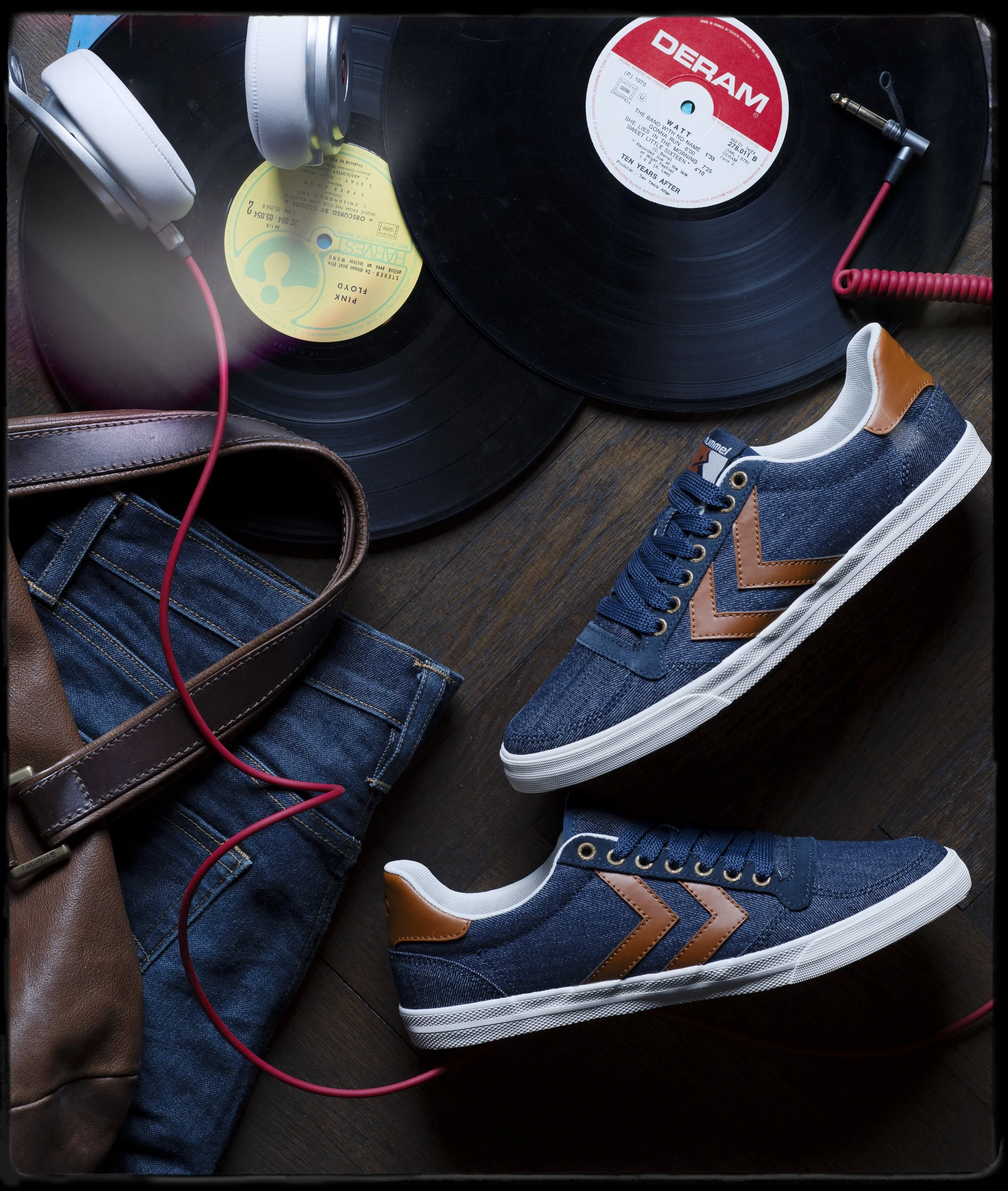 Platine vinyle avec deux disques, casque audio gris, chaussures de sport bleues et blanches, sac en cuir marron, jeans et câble audio rouge.