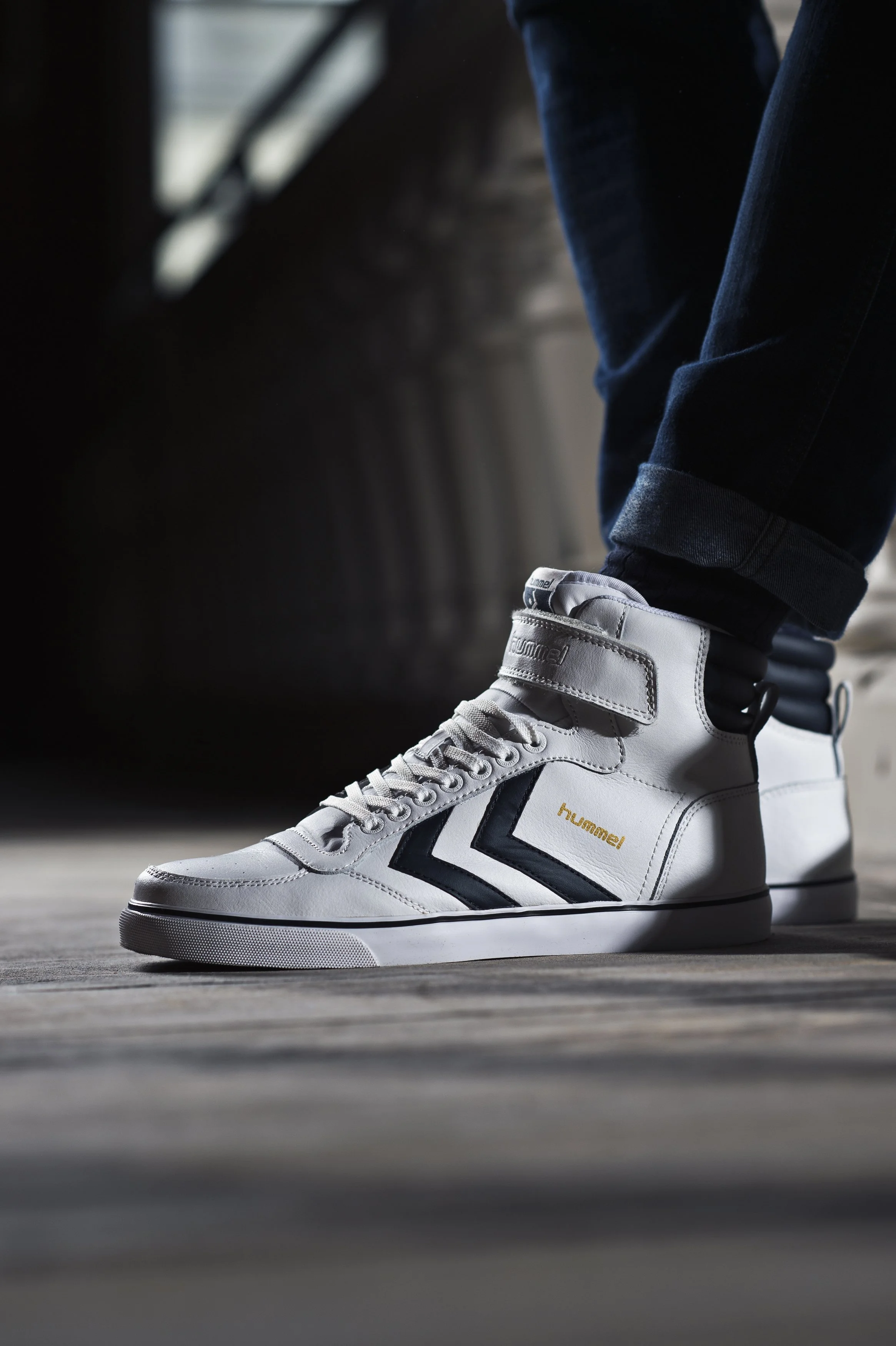 Chaussures blanches de sport avec la marque Hummel, portées par une personne en jean, dans un environnement intérieur avec un sol en bois.