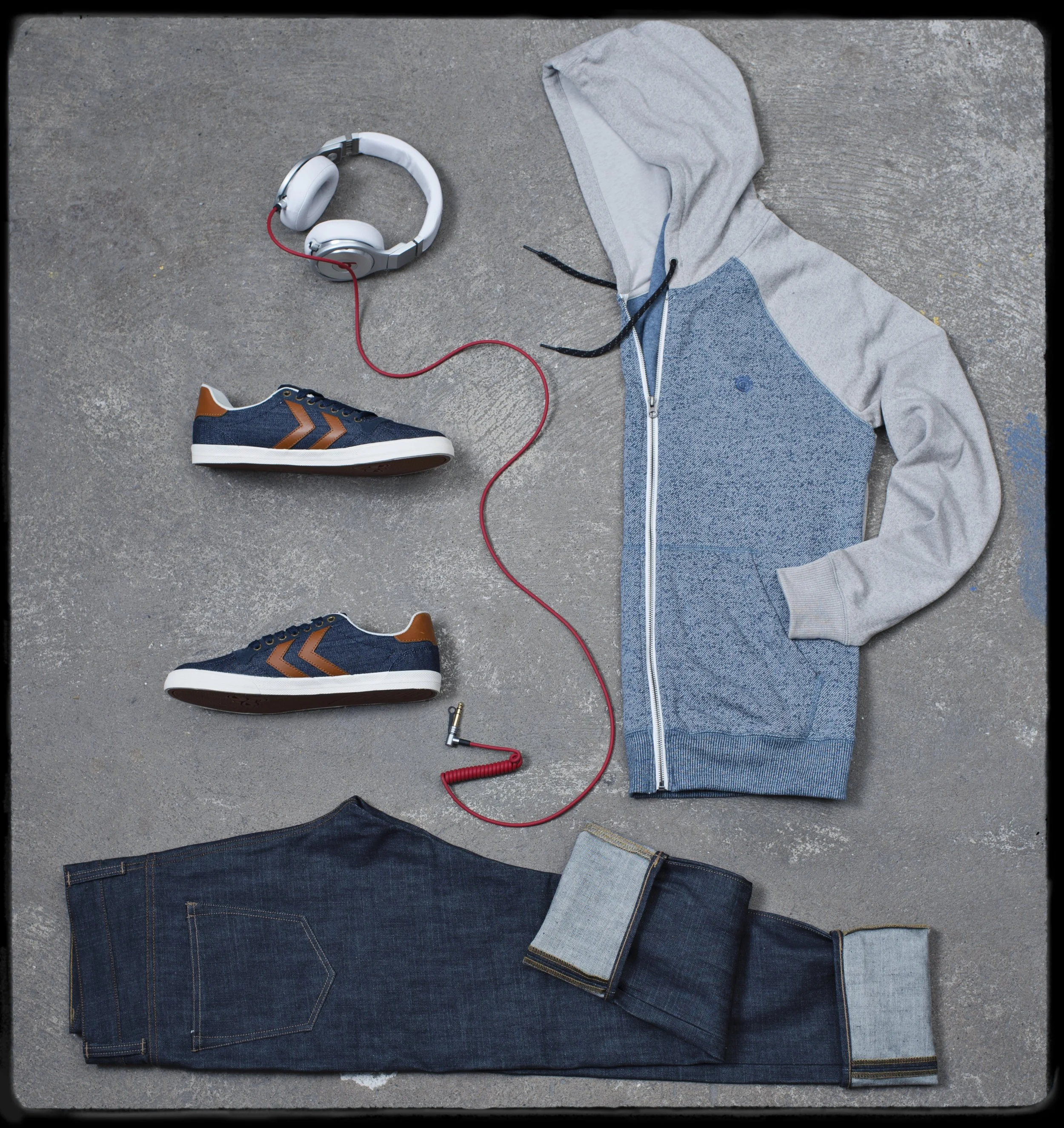 Une paire de chaussures de sport, un sweat à capuche gris, un jean, et un casque audio avec un câble rouge, tous disposés à plat sur un sol en béton.