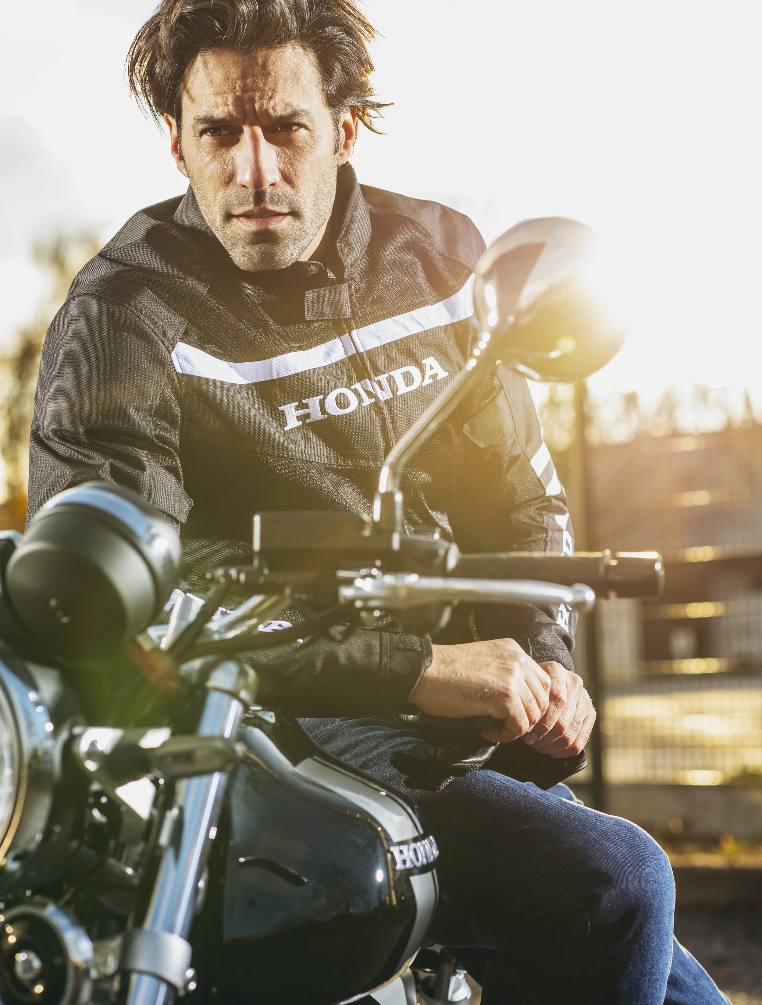 Un homme en tenue de motard sur une moto Honda, regardant vers la gauche avec un fond lumineux en fin de journée.