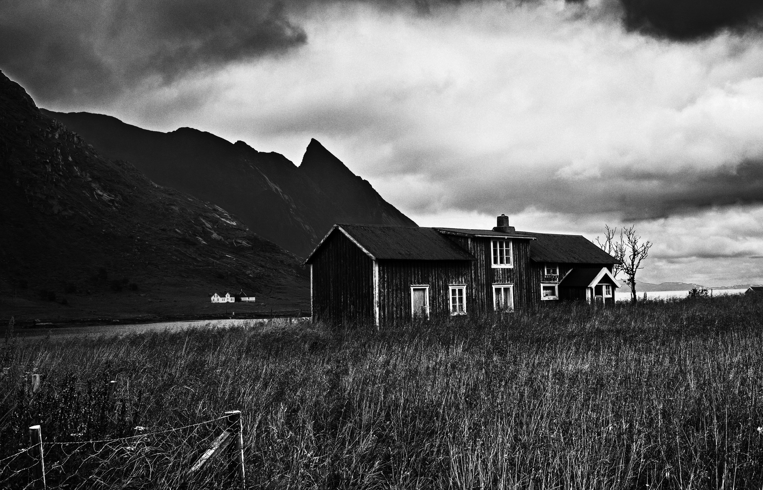 Une maison en bois dans un paysage rural avec des montagnes en arrière-plan, ciel nuageux, en noir et blanc.