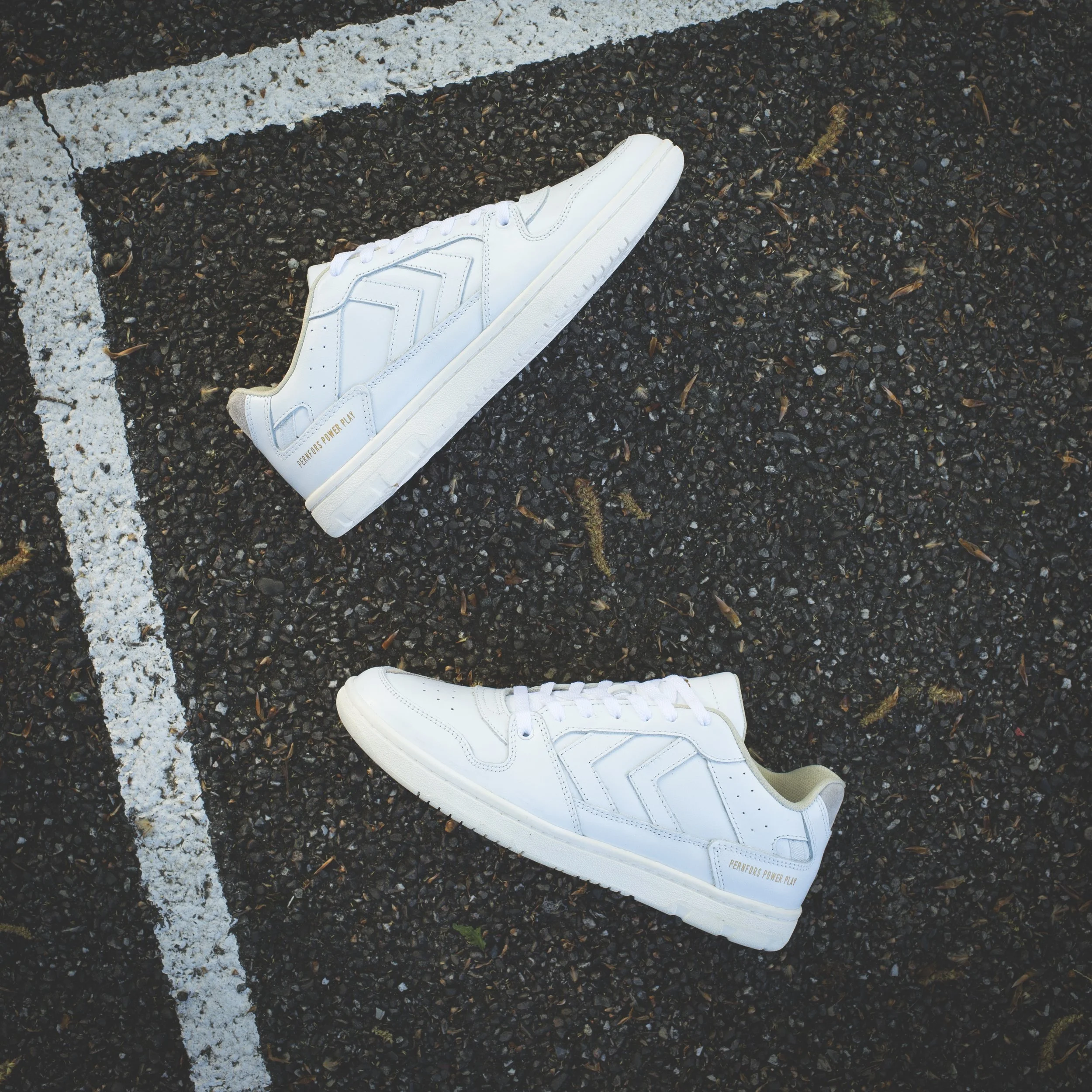 Une sneaker blanche posée sur une route goudronnée, près de la ligne blanche peinturée, vue du dessus - marque HUMMEL