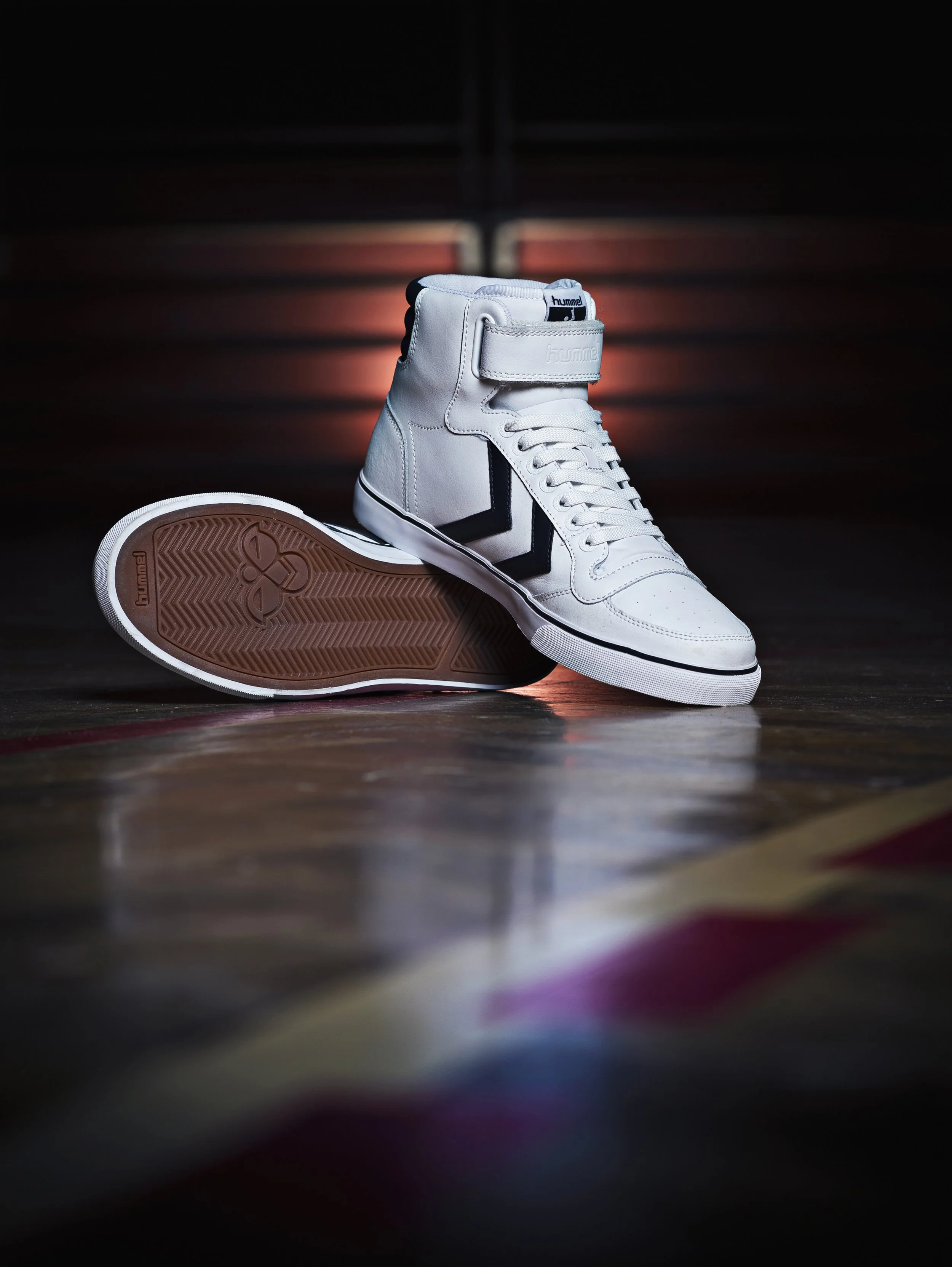 Une chaussure de basket blanche avec des détails noirs, posée sur un sol en bois; l'image met en valeur la semelle et le côté de la chaussure, avec un arrière-plan sombre.