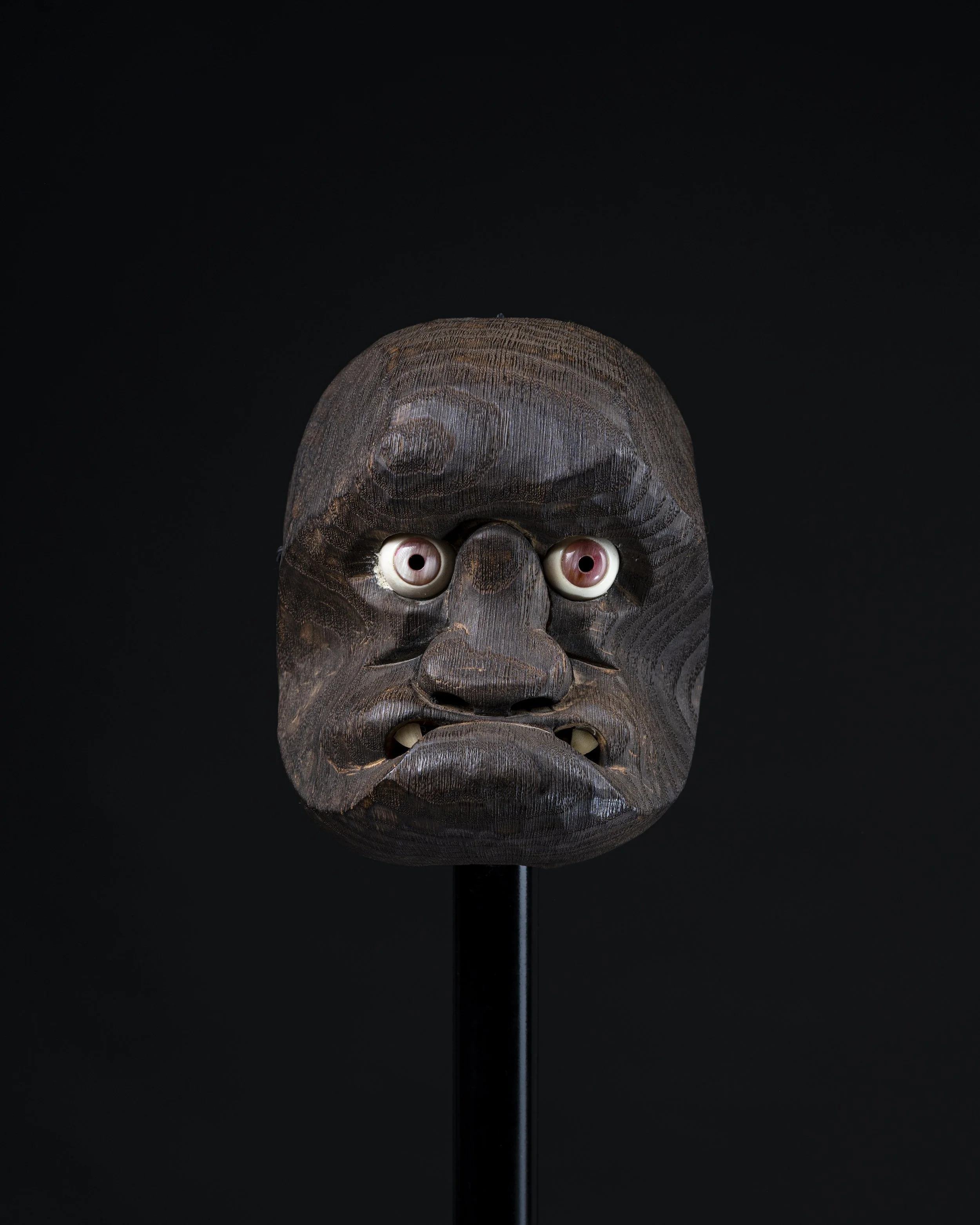 Une sculpture en bois représentant une tête avec des yeux globuleux en porcelaine, fixée sur un support noir.