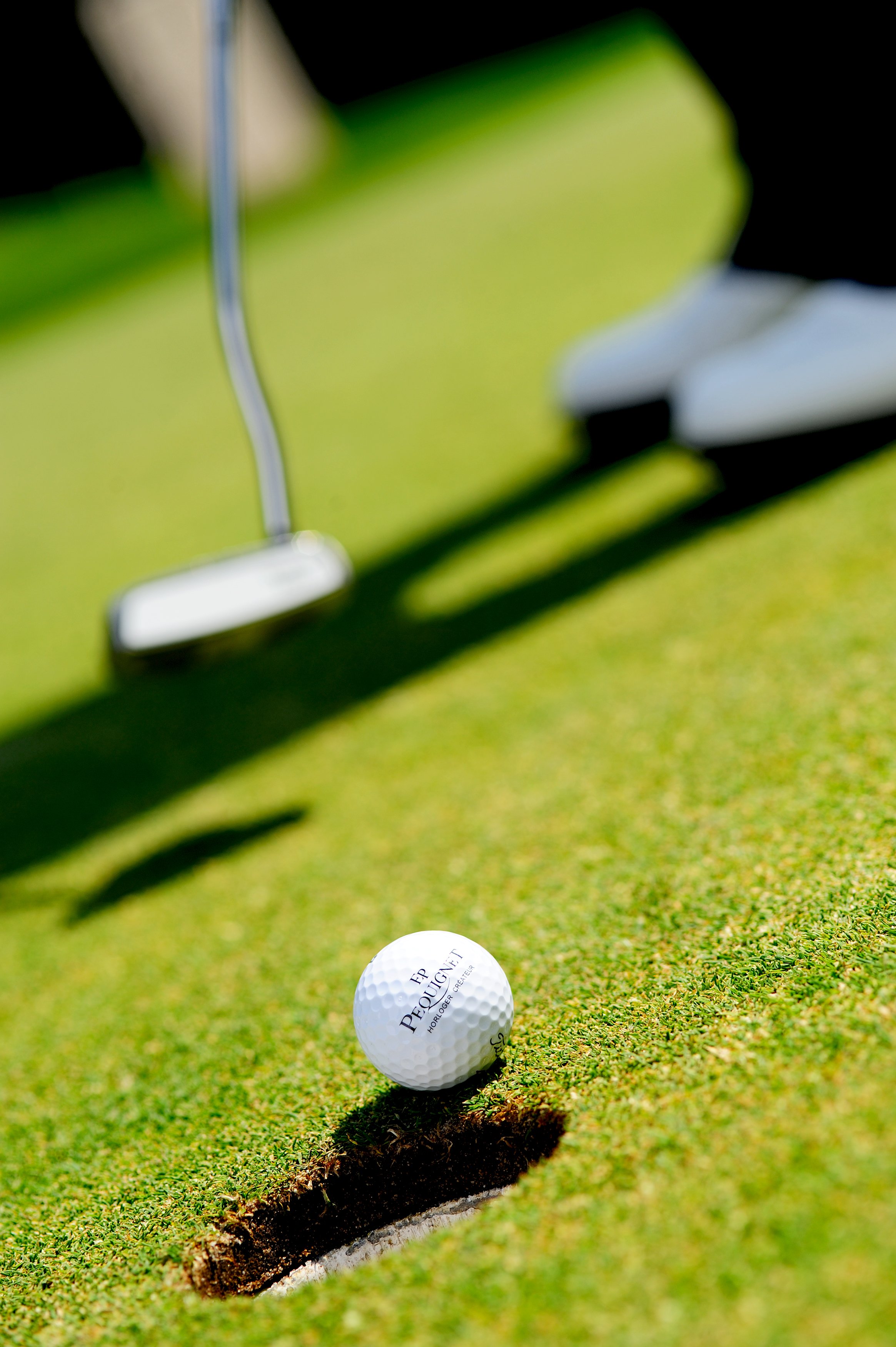 Une balle de golf blanche portant la marque "PEQUIGNET" repose près d'une tranche de trou sur un green de golf vert. Un club de golf est visible en arrière-plan, prêt à frapper la balle.