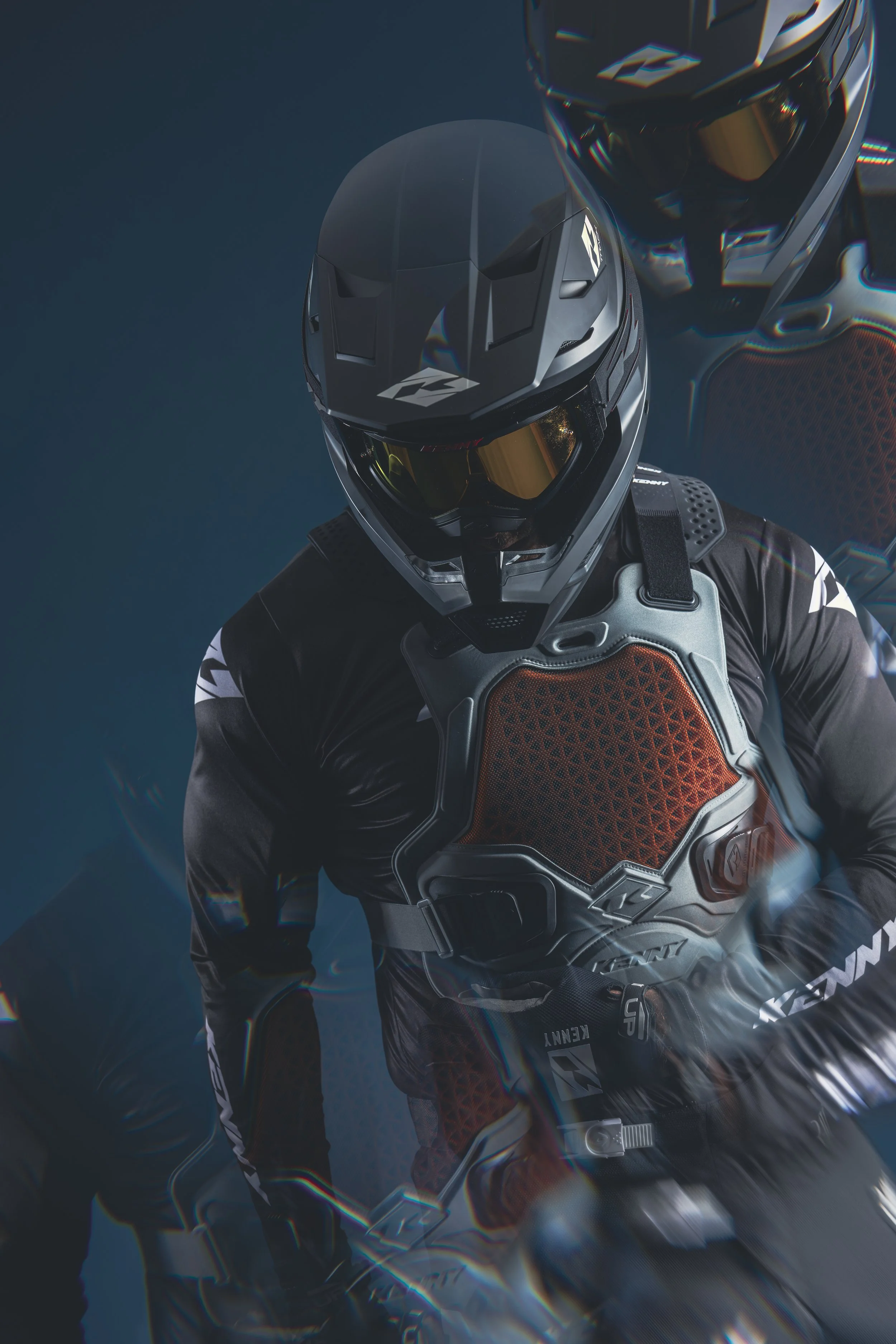 Un pilote de motocross portant un casque noir avec des visières dorées et une tenue de protection noire et orange, sur fond sombre.