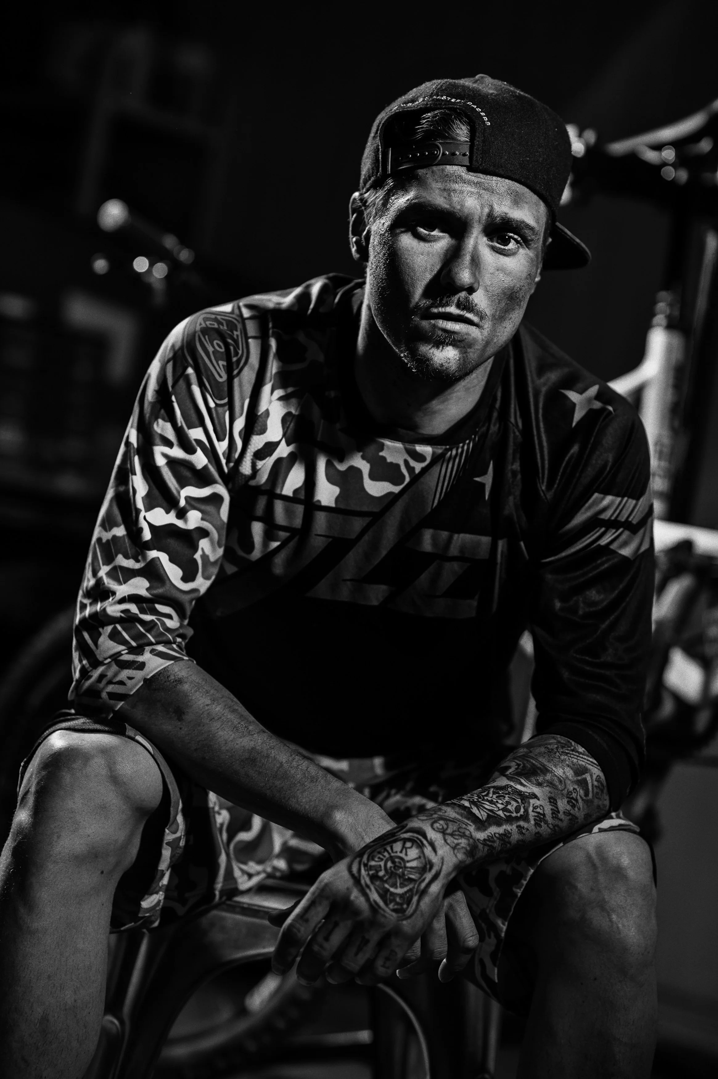 Homme jeune avec une casquette à l'envers, tatoué, portant un maillot de sport, en posture assise dans un environnement sombre, photographie en noir et blanc.