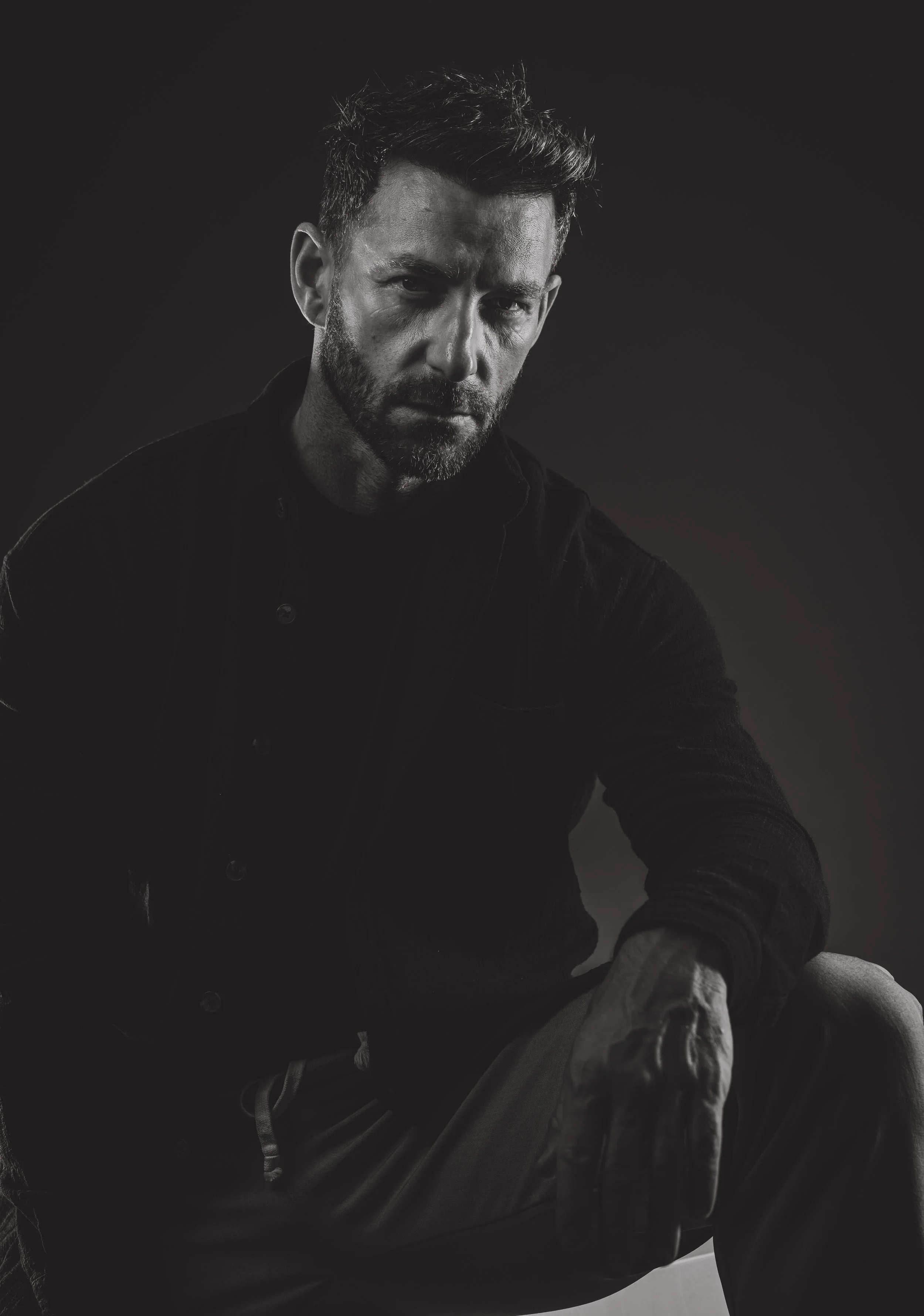 Un homme à barbe porte une veste noire et regarde sérieusement vers l'objectif, dans un contexte sombre et monochrome.