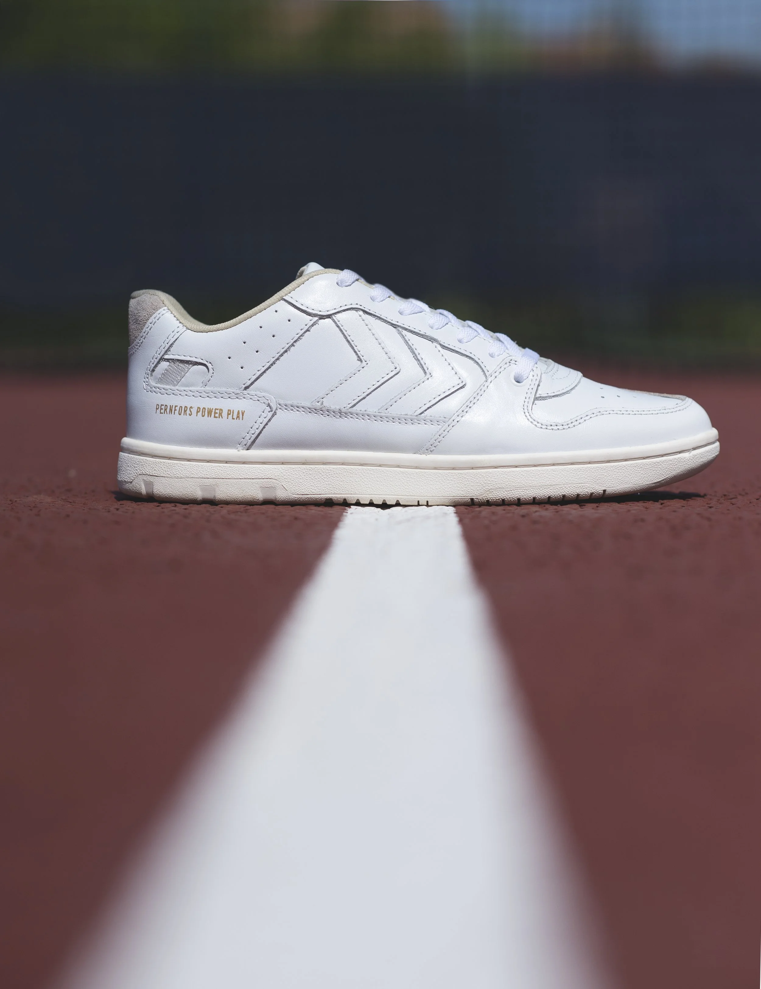 Chaussure blanche pour le sport posée sur une piste d'athlétisme. texture en cuir, détails en relief, motif en zigzag, inscription 'PERNFORS POWER PLAY' sur le côté - marque HUMMEL