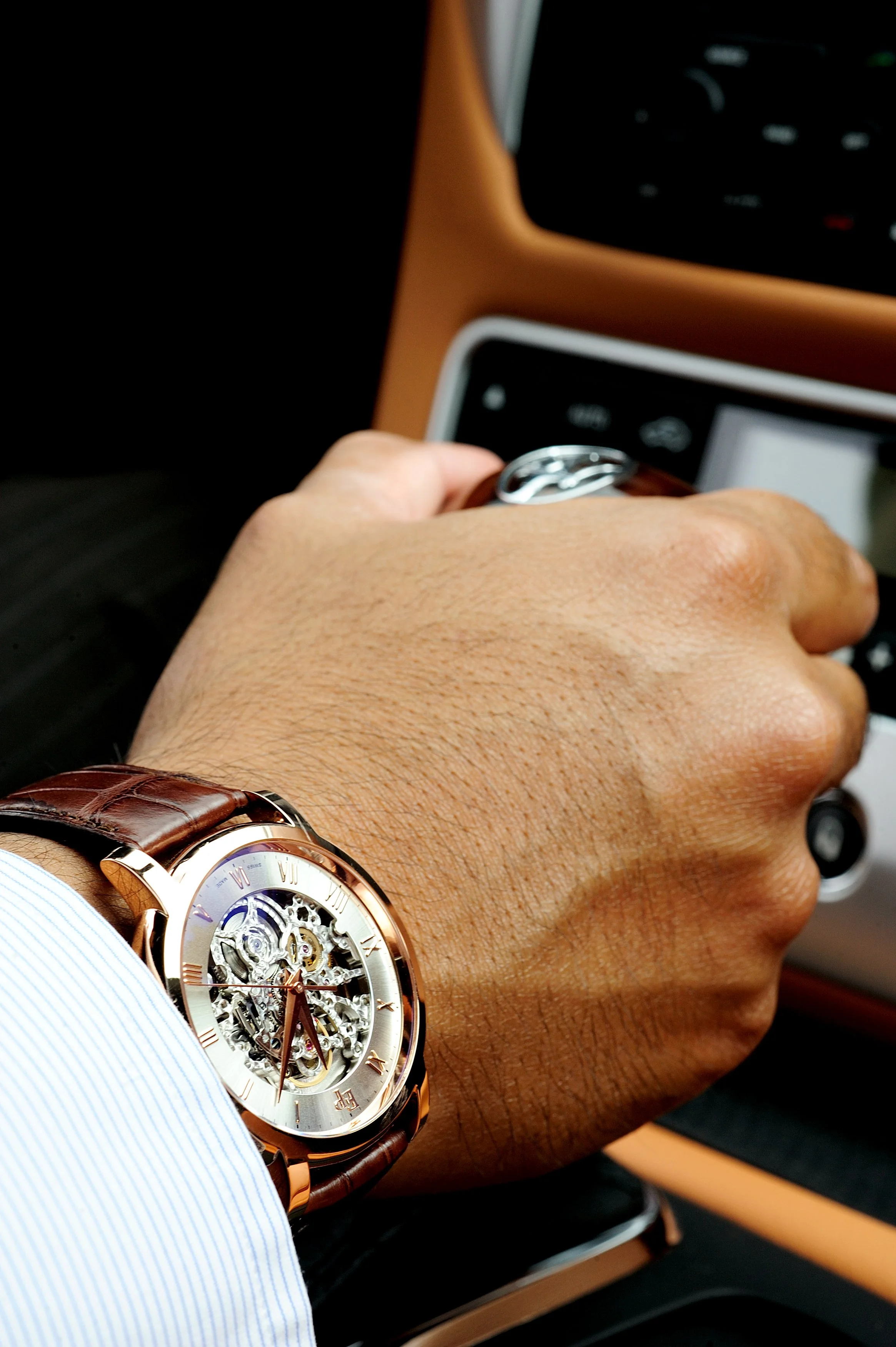 Poignet d'une personne portant une montre en or rose avec un cadran ouvert, montrant le mécanisme intérieur, vêtu d'une chemise à rayures bleues, à l'intérieur d'une voiture.