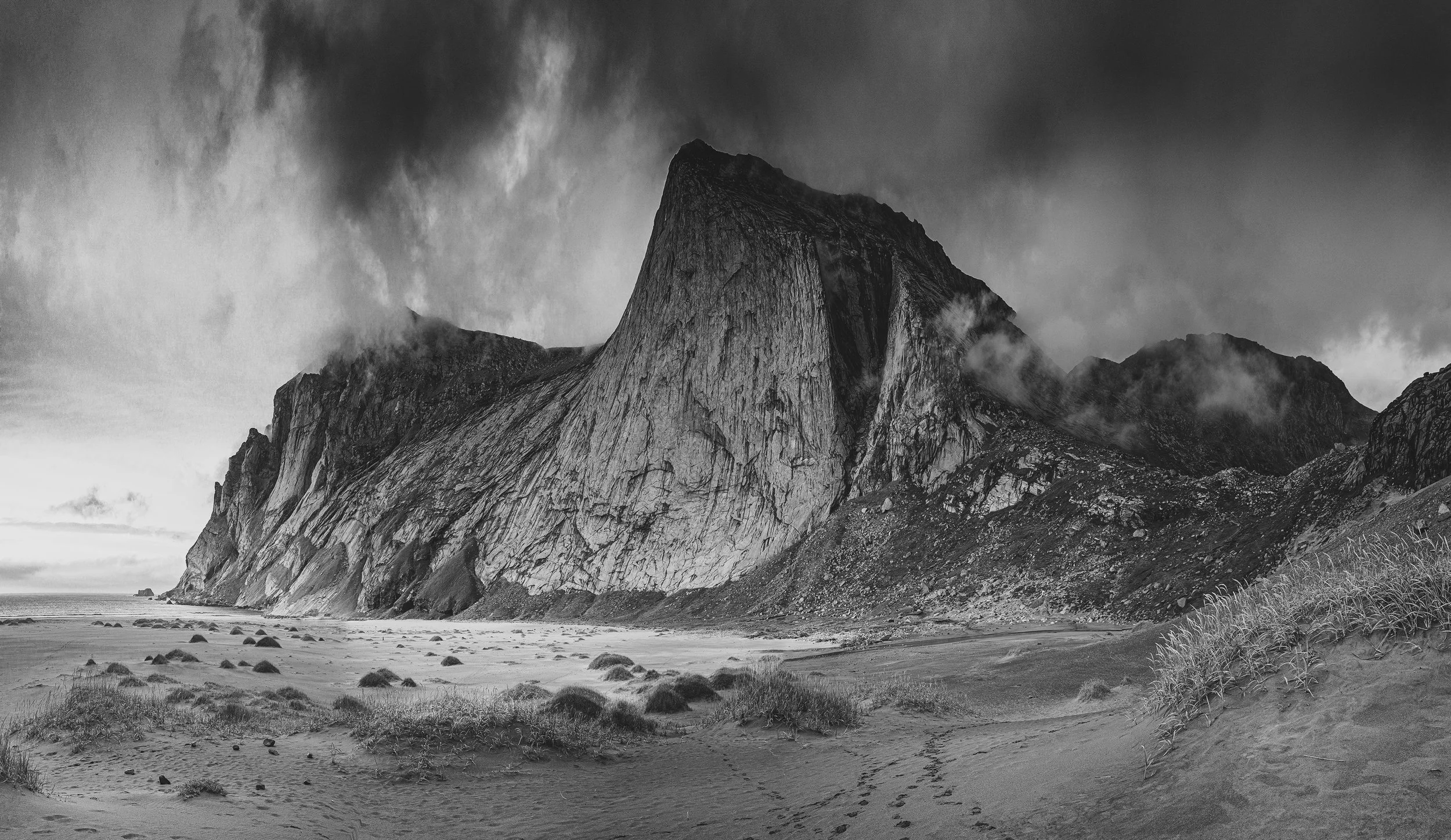 Montagne rocheuse en noir et blanc avec ciel nuageux, plage et végétation au premier plan.