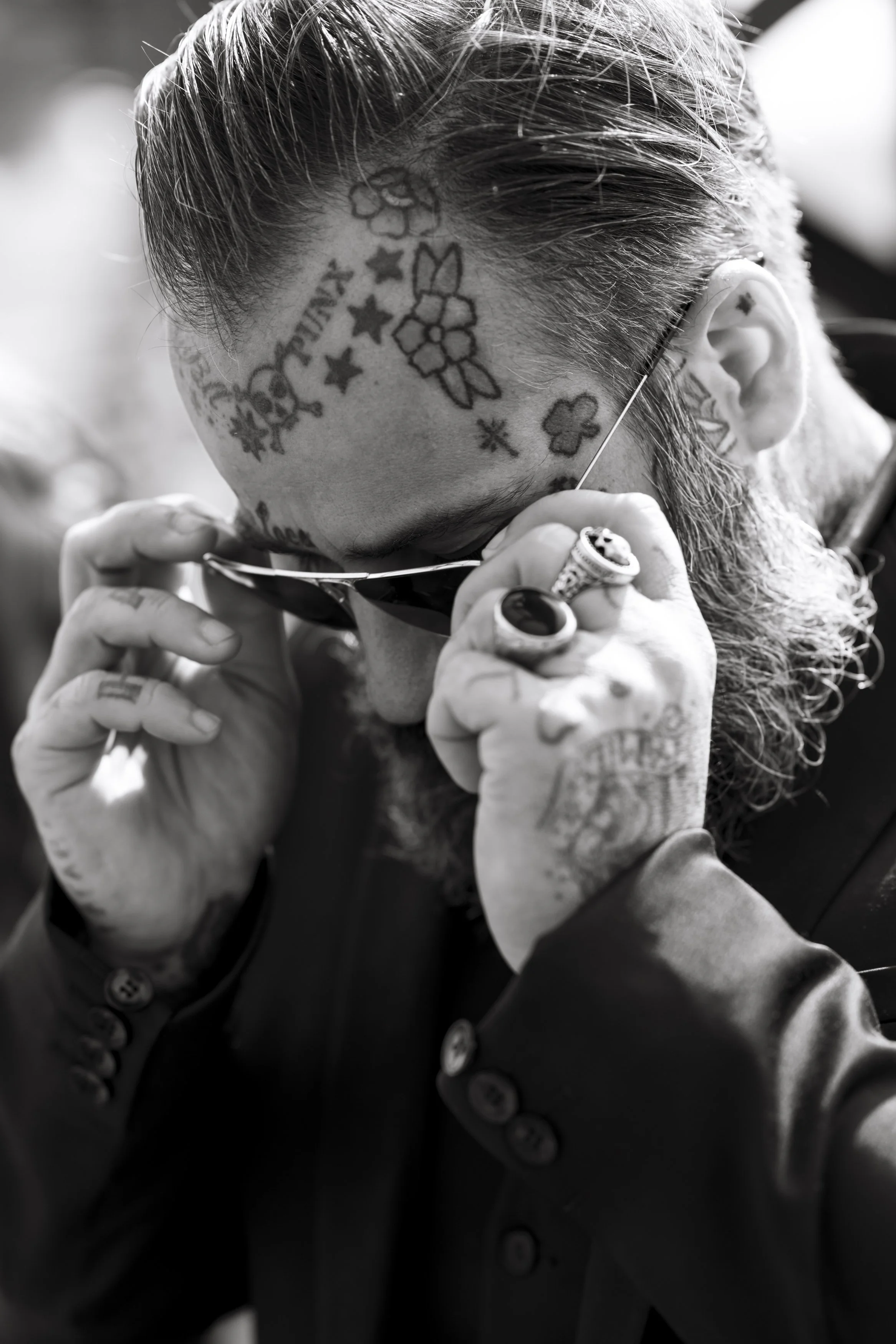 Un homme avec des tatouages sur le visage et la tête, portant des lunettes et ajustant celles-ci, vêtu d'une veste boutonnée. La photo est en noir et blanc.
