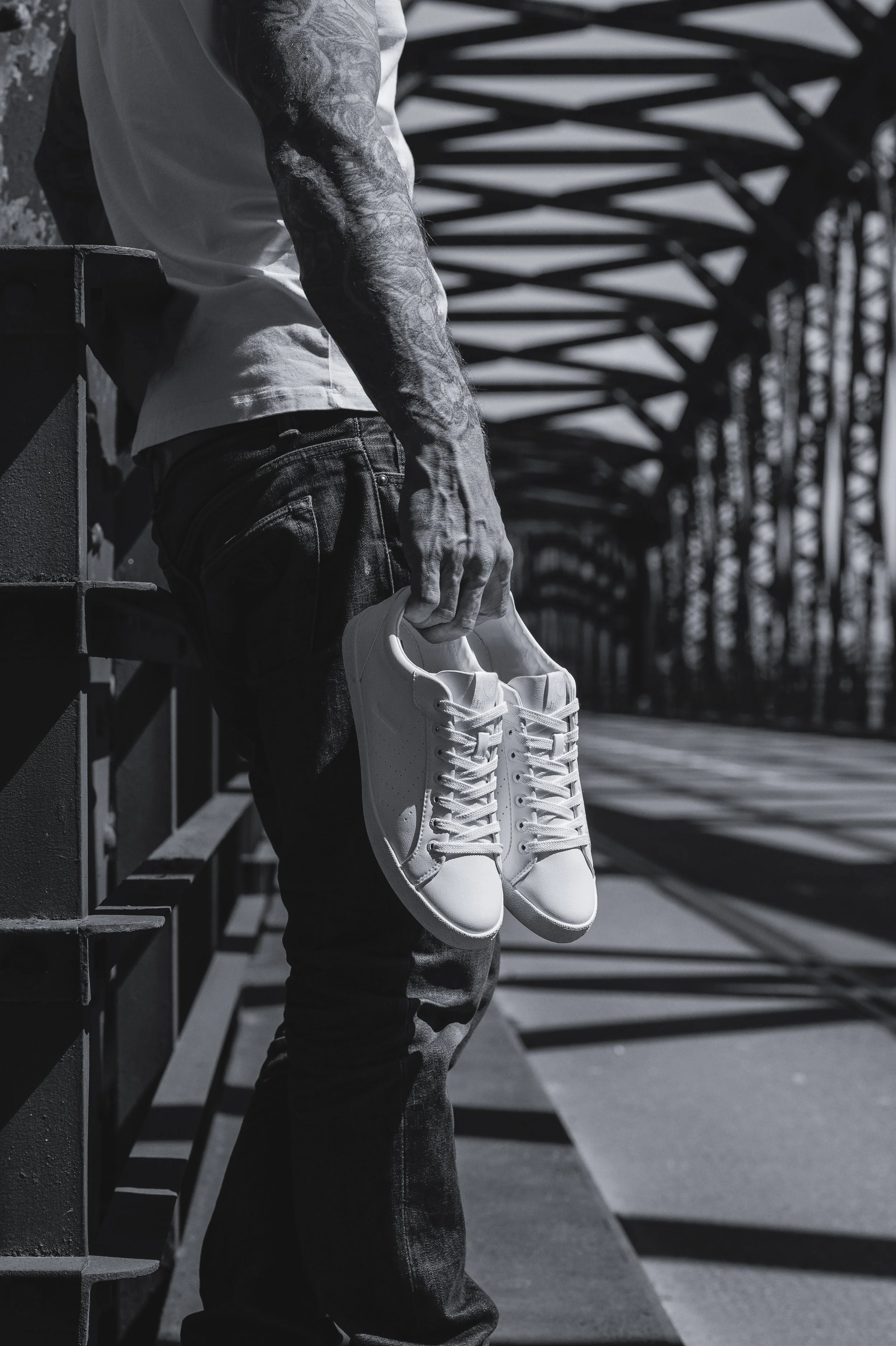 Un homme tenant une paire de baskets blanches contre une rambarde en métal dans un environnement urbain en plein air, avec un pont métallique en arrière-plan.