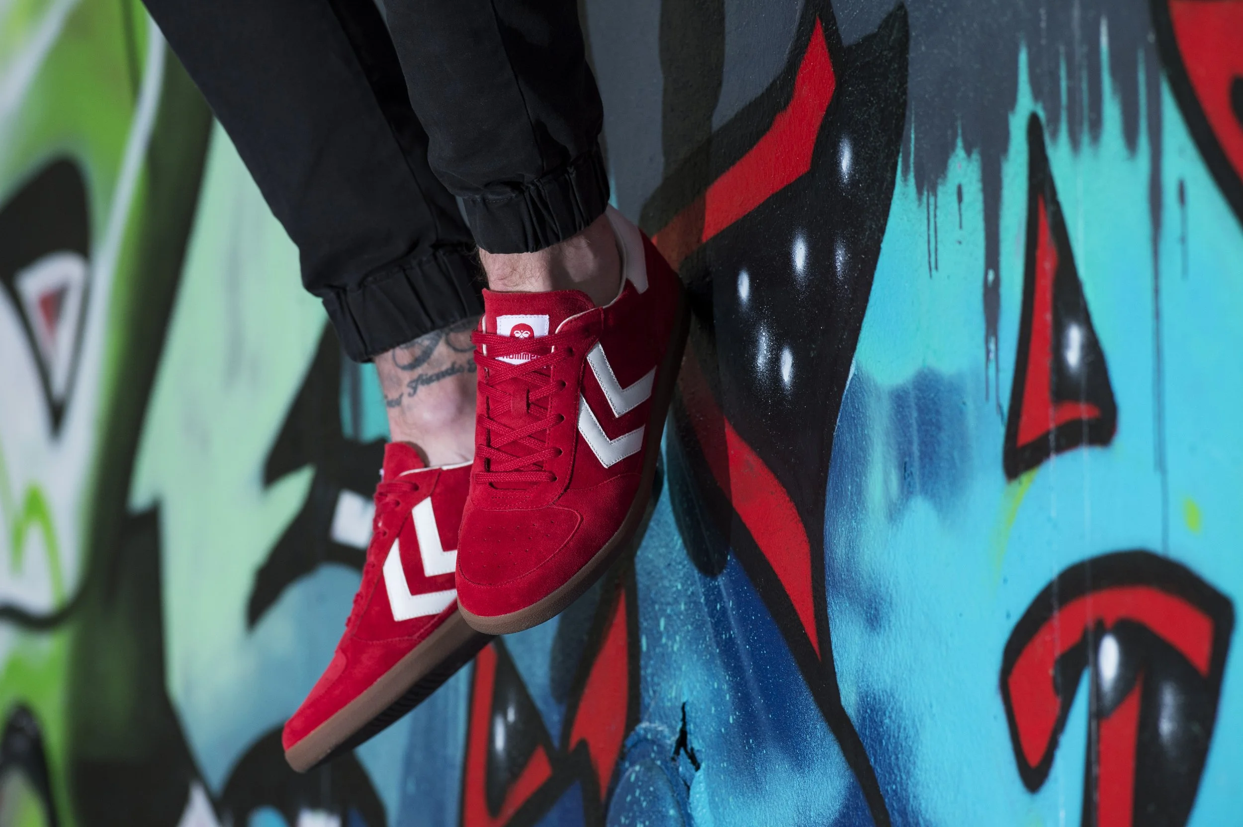 Une personne portant des chaussures de sport rouges avec des détails blancs, posée sur un mur de graffiti coloré.