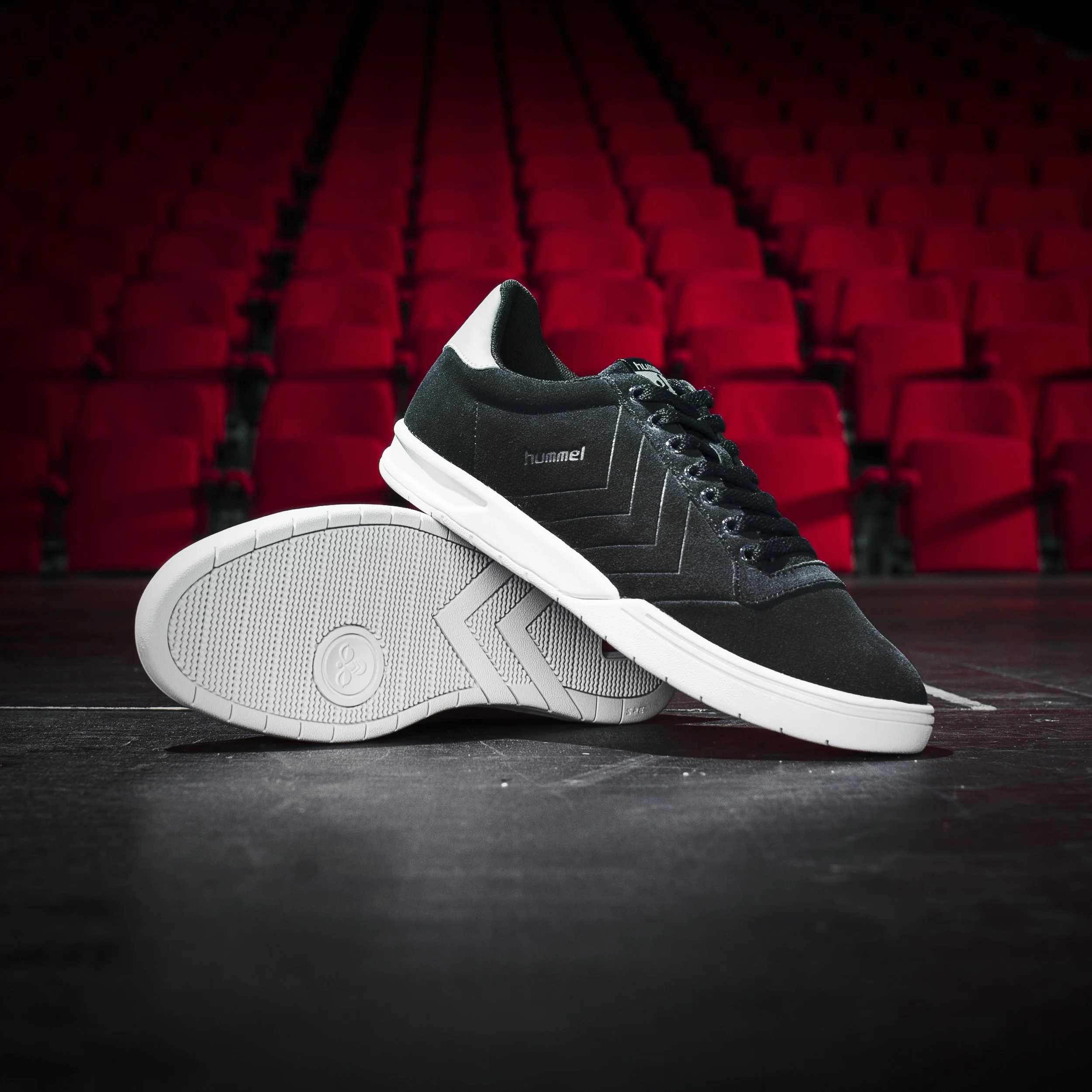 Une chaussure de sport noire et blanche posée sur un sol sombre dans une salle de spectacle ou cinéma, avec des sièges rouges en arrière-plan.