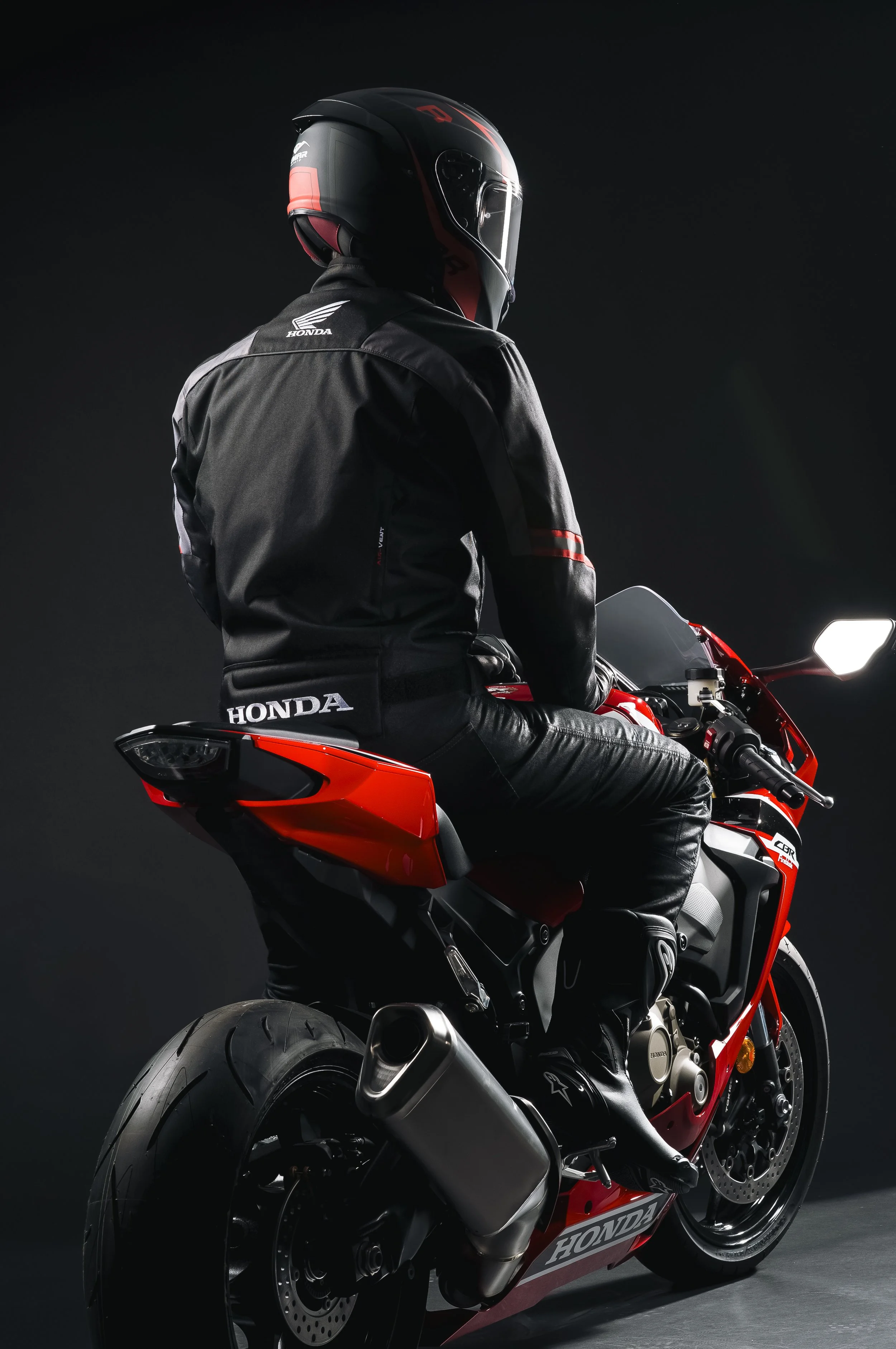 Un homme portant un casque intégral et une veste noire avec le logo Honda, assis sur une moto rouge et noire, contre un fond sombre.