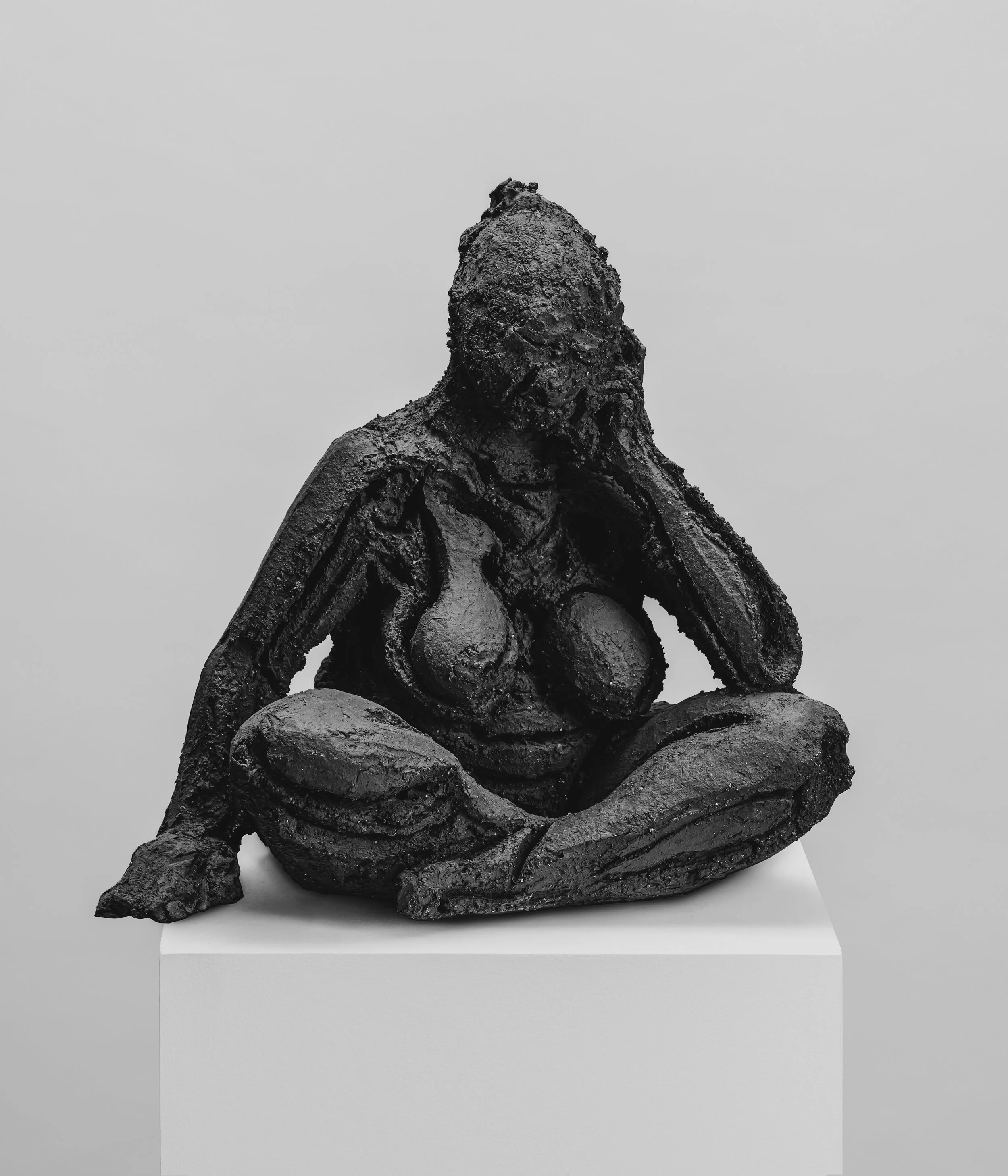 Sculpture en bronze représentant une figure humaine en position agenouillée, avec une tête pointue et des bras repliés, posée sur un socle blanc.