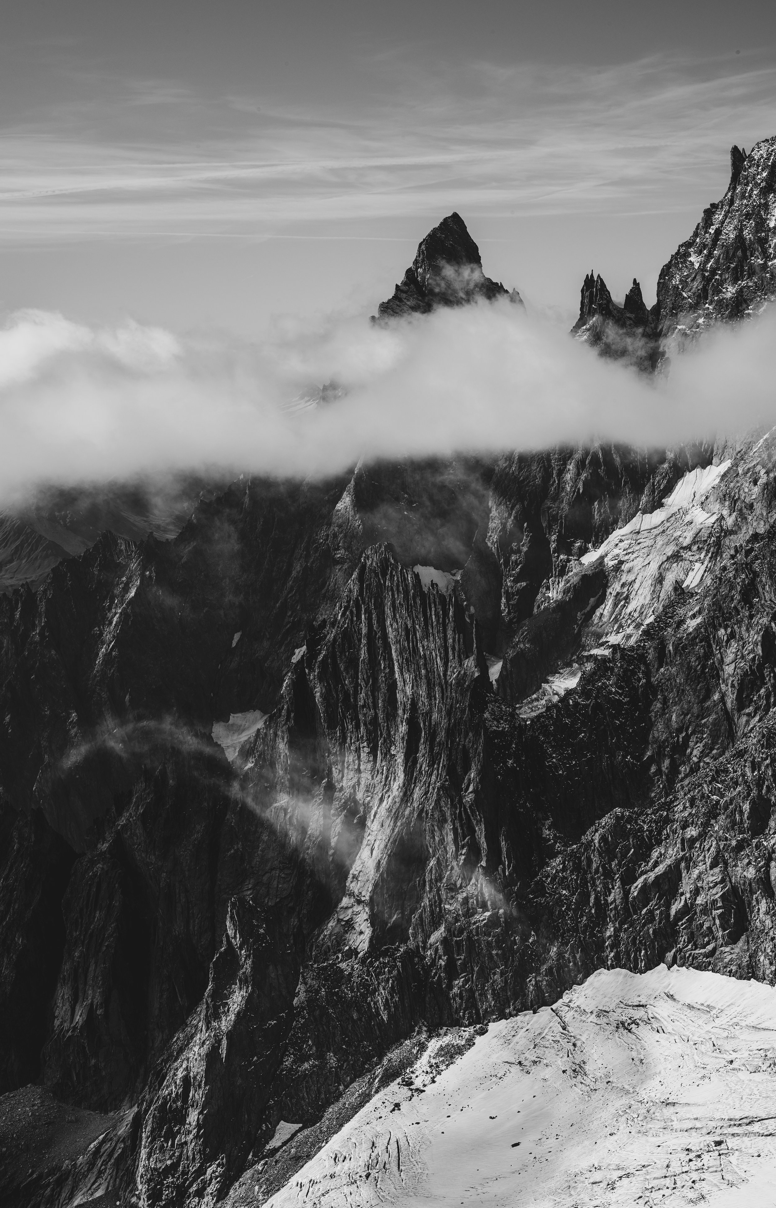 Montagnes rocheuses enneigées partiellement enveloppées de nuages, vue en noir et blanc.
