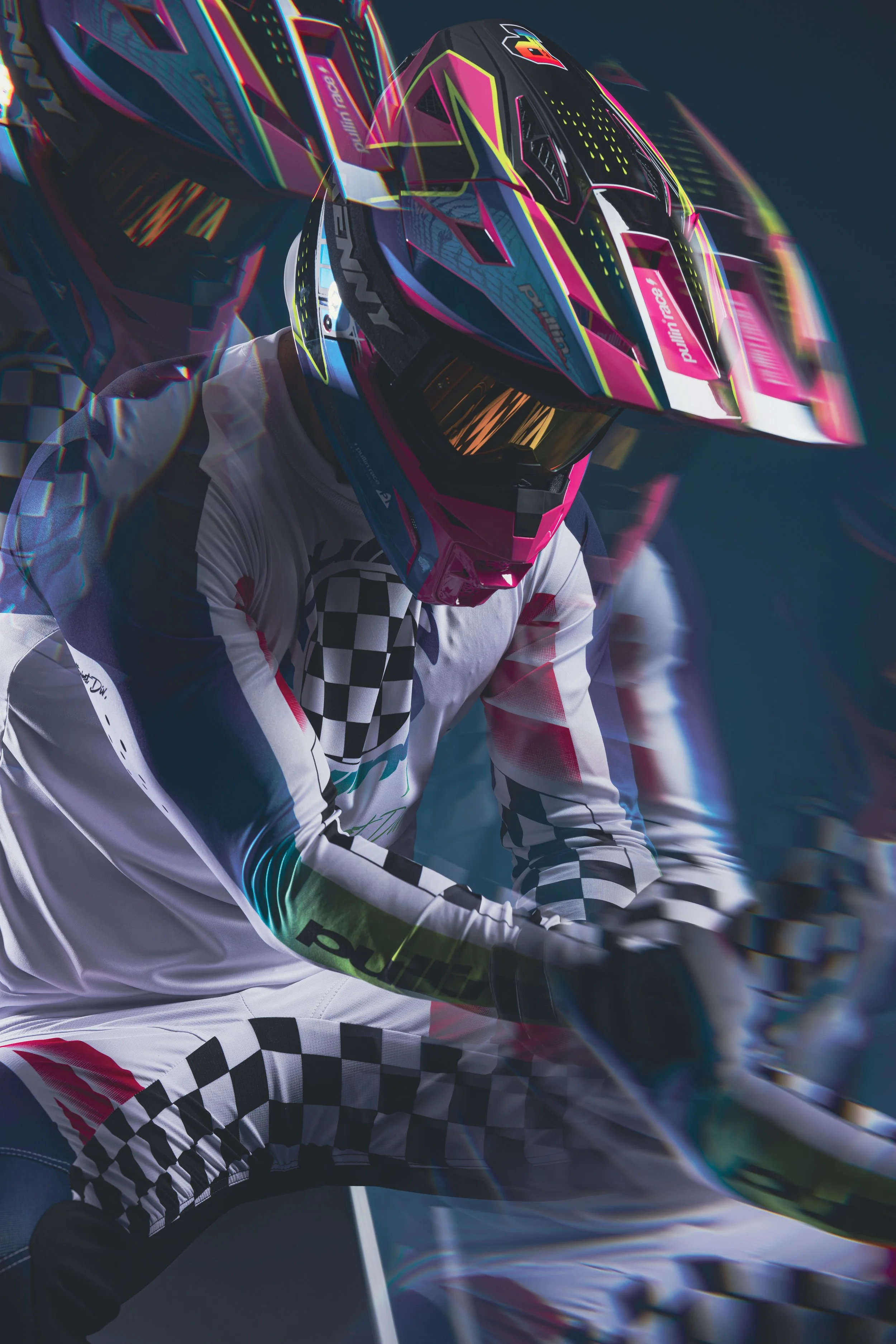 Un pilote de course portant un casque de couleur vive, un costume de course à motifs noirs et blancs, en position sur une moto de course, avec un fond sombre et un effet de mouvement.